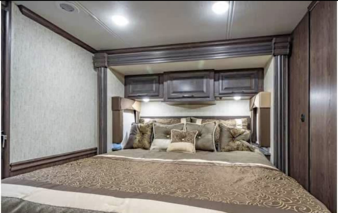 Luxurious RV rental bedroom featuring a large bed, decorative pillows, overhead cabinets, and rich wood accents.. Coachmen Sportscoach 2019