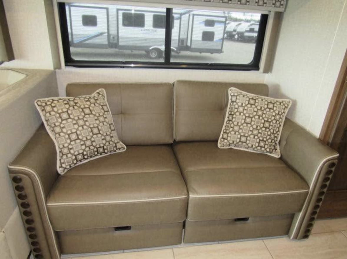 RV rental interior: cozy sofa with storage, decorative pillows, and a window view of other trailers.. Coachmen Sportscoach 2019