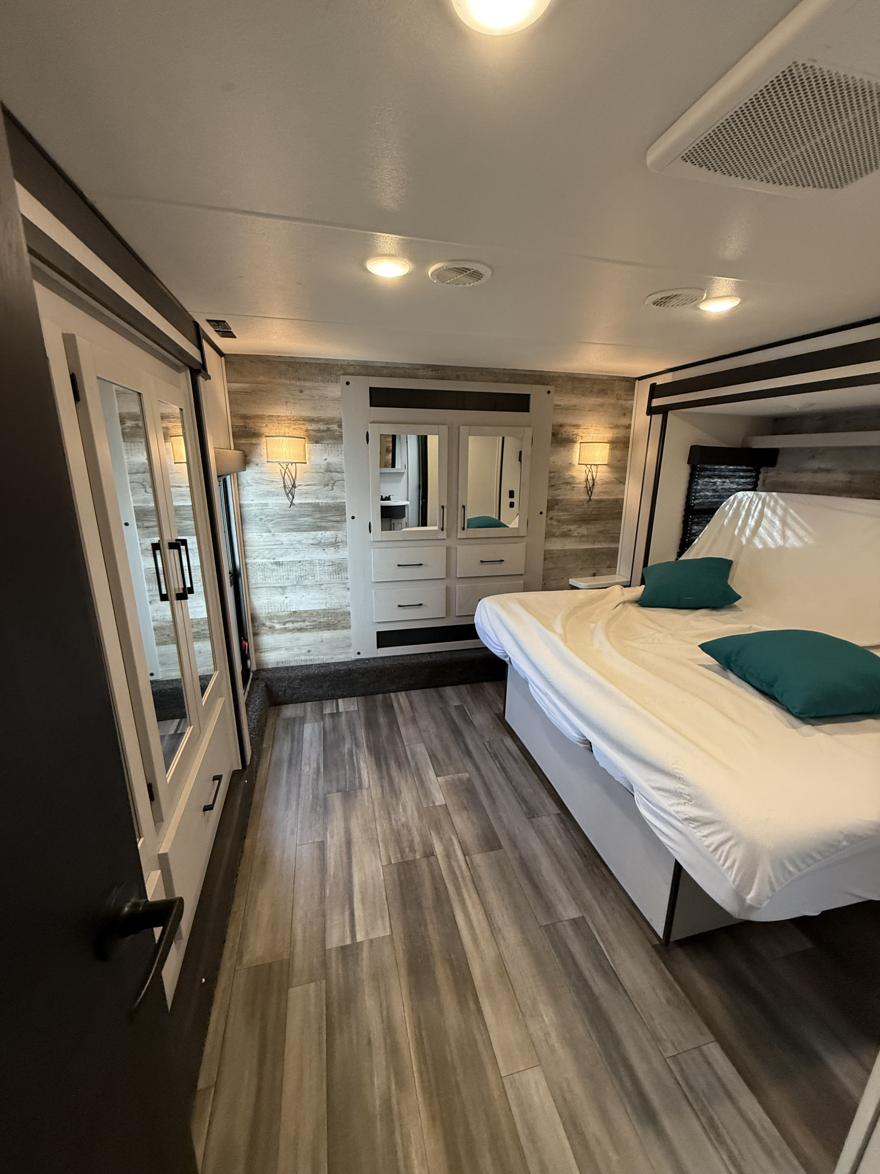 Cozy RV rental bedroom featuring wood floors, a white bed, paneled walls, a mirrored dresser, and a sliding door.. Heartland RVs Torque Toy Hauler 2024