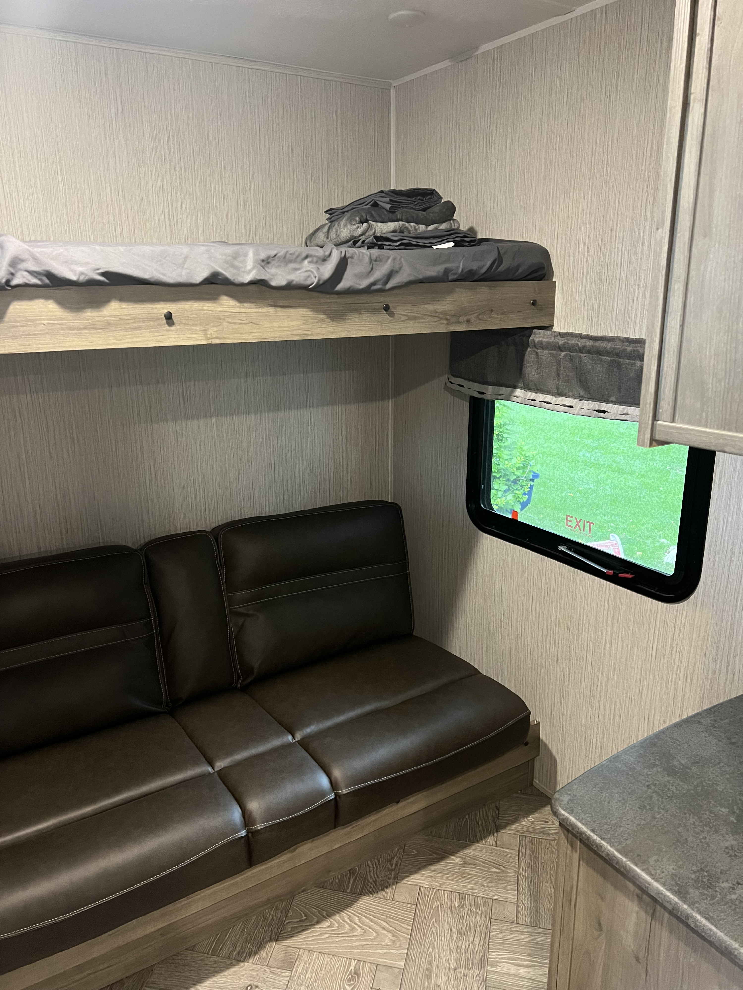 RV rental interior: cozy bunk bed with grey bedding, brown couch, and window showing an "EXIT" sign & green view.. Palomino Puma 2022