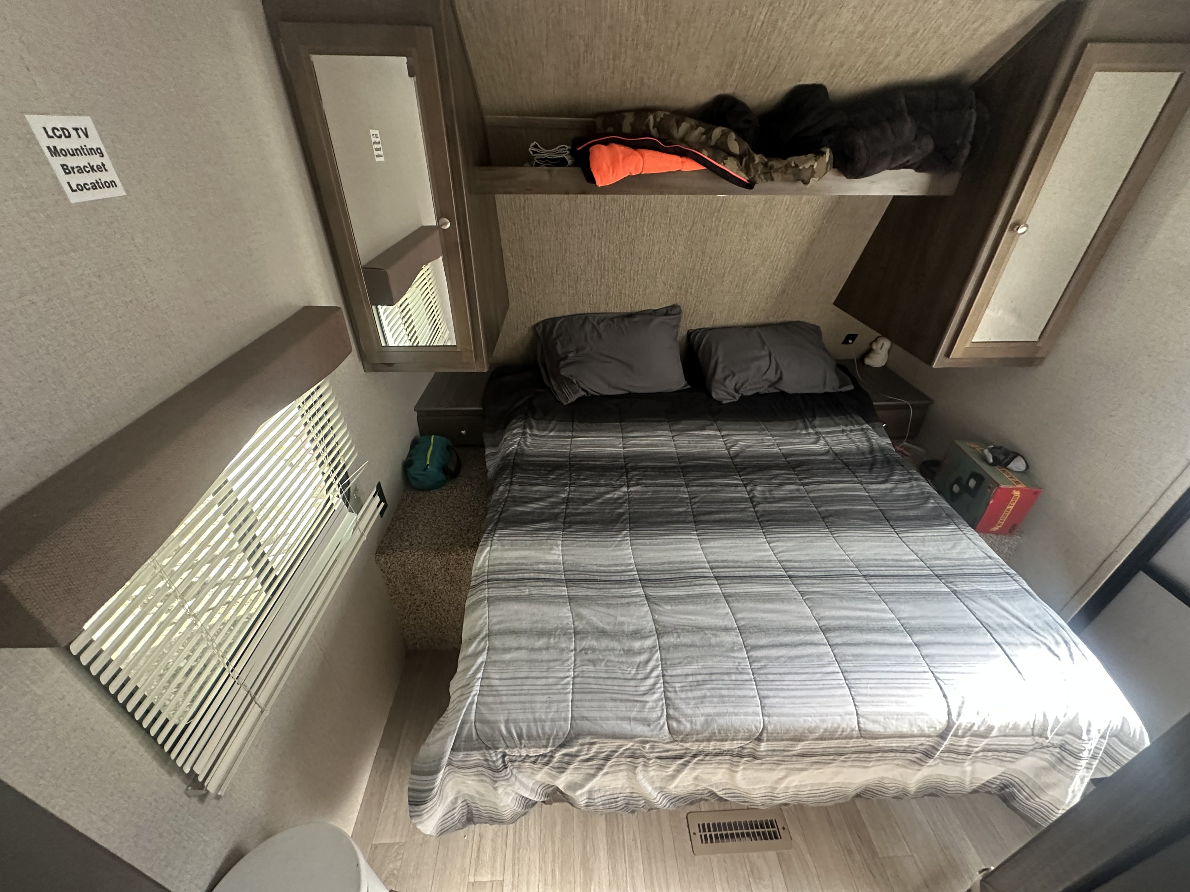 Cozy RV rental bedroom featuring a large bed, overhead storage, window with blinds, and a TV mounting sign.. Dutchmen Coleman Lantern 2019