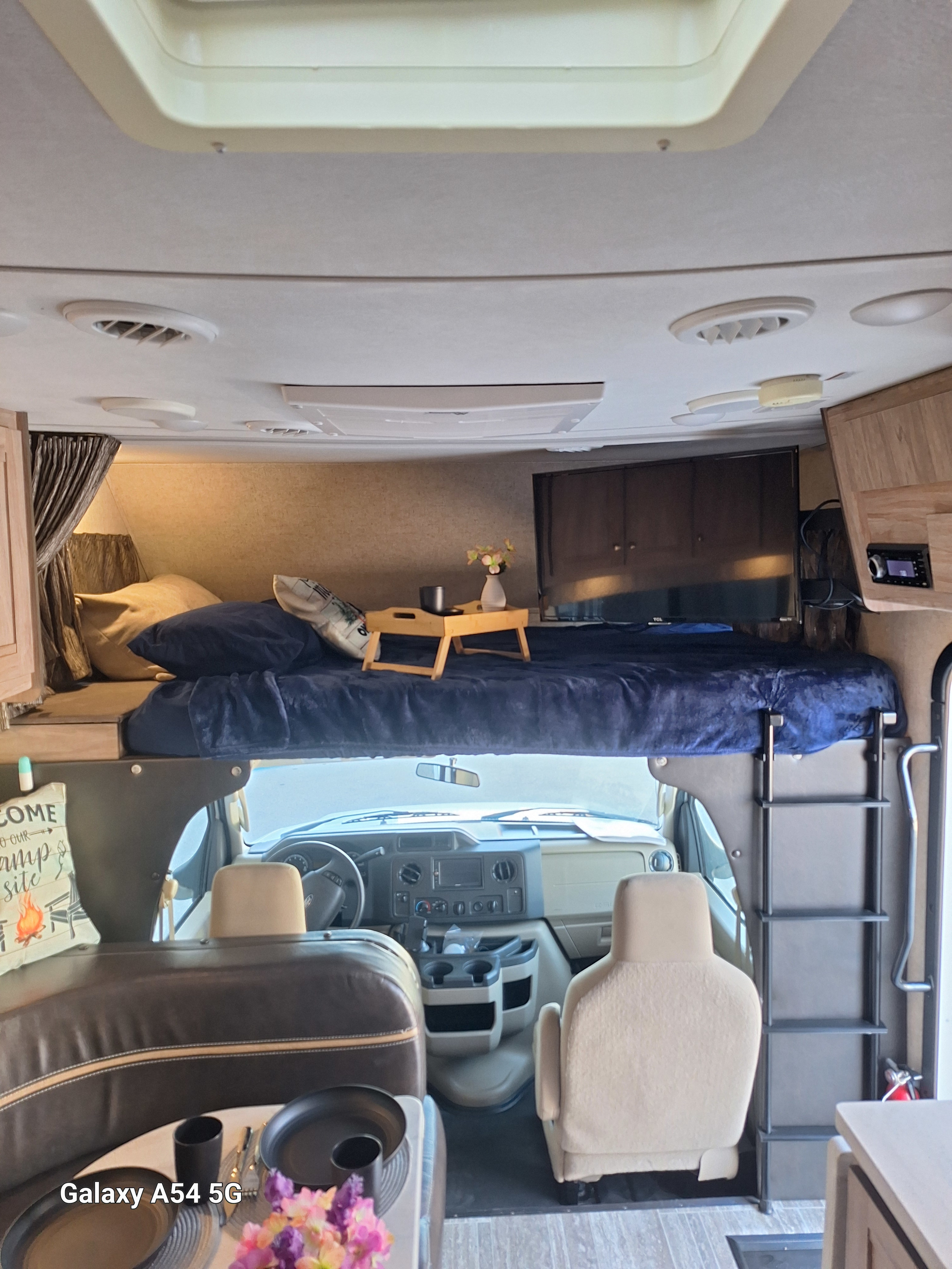 This cozy RV rental interior features an overhead bed with blue bedding, a dining area, and a driver's cockpit.. Forest River Sunseeker LE 2018