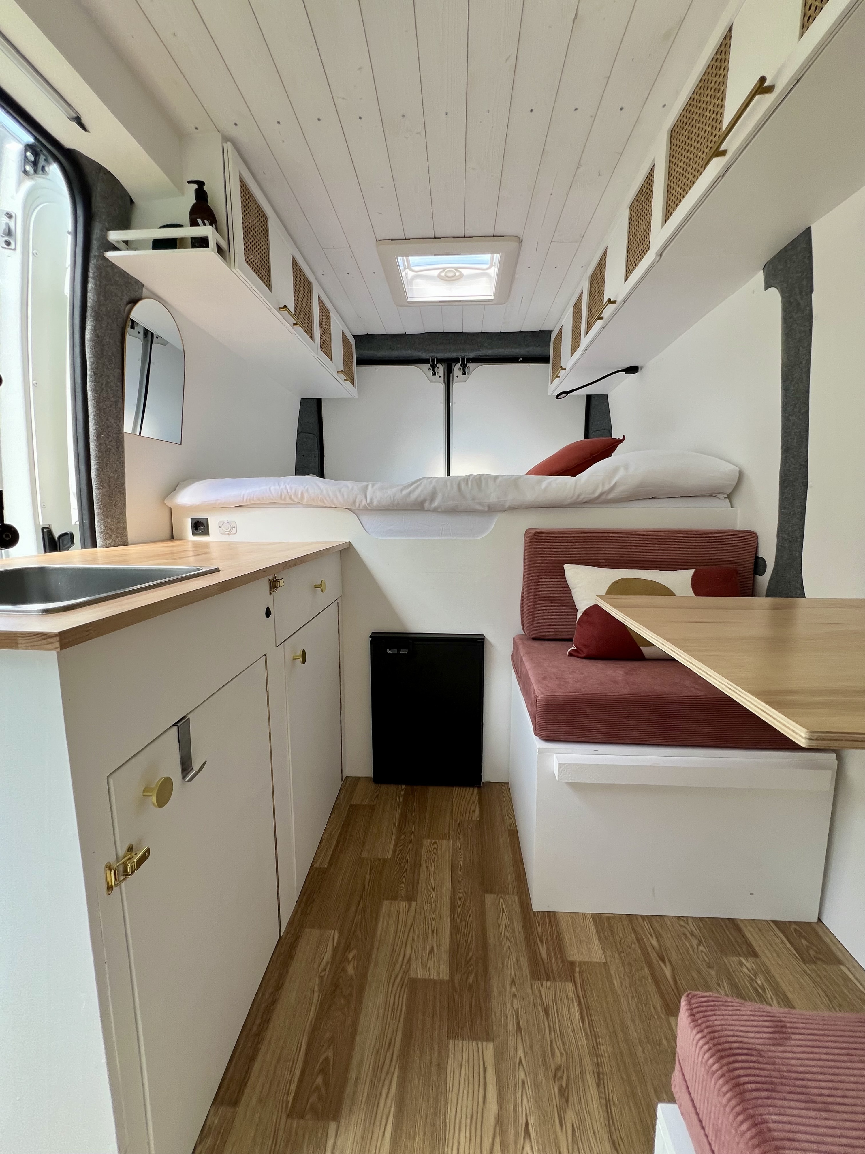 View of a bright, modern RV rental interior with a wood-planked ceiling, white cabinets, kitchen, bed, and dining area.. Peugeot Boxer 2021