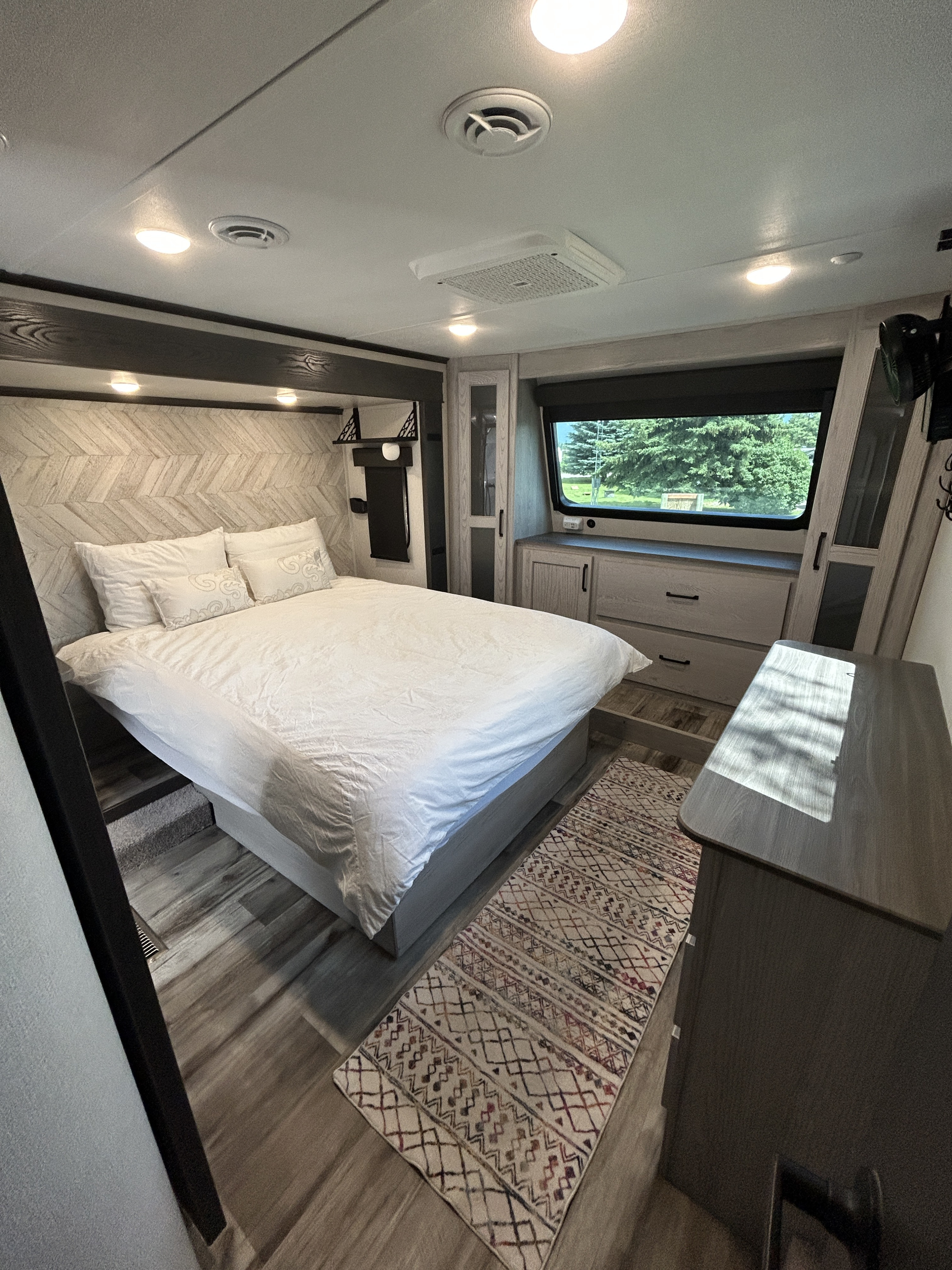 Bright RV rental bedroom showcasing a white bed, geometric headboard, ample storage cabinets, and a window with a treeline view.. Keystone RV Montana High Country 2023