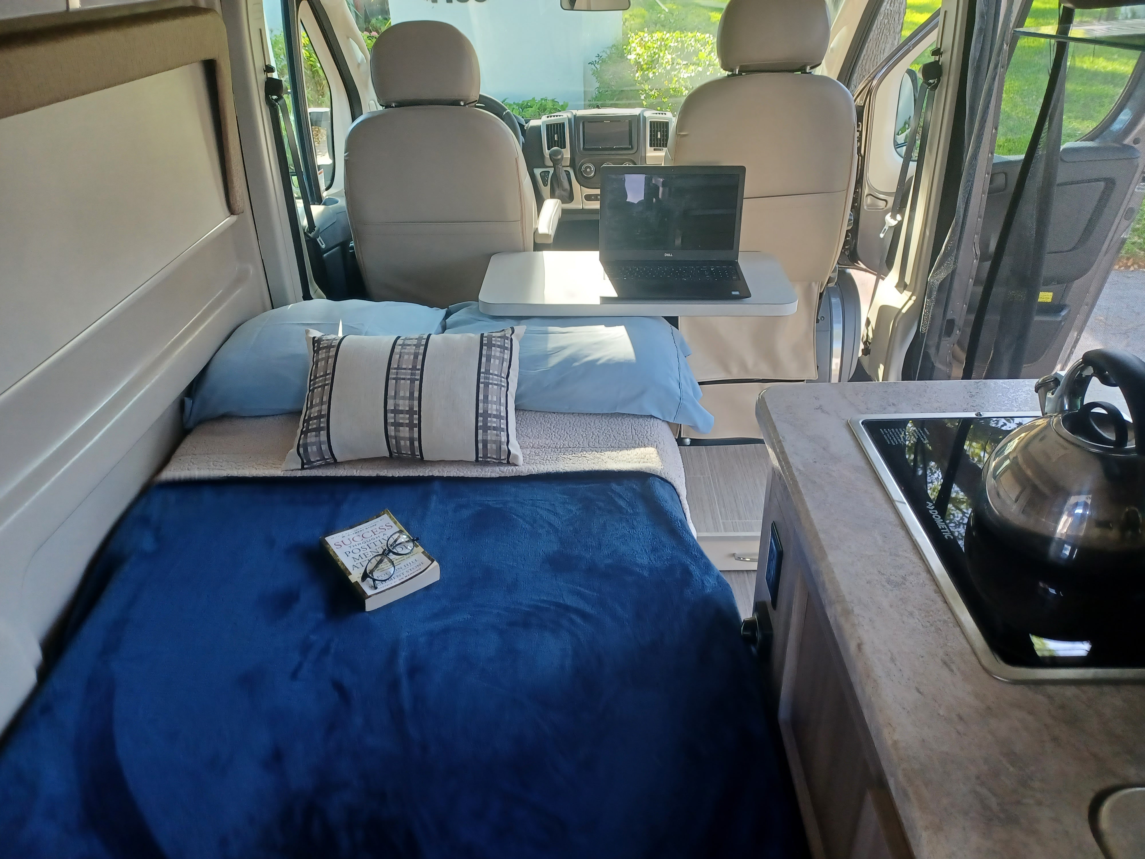Inside a comfy RV rental: plush bed with book, compact workstation with laptop, & a functional kitchenette.. Thor Scope 2022