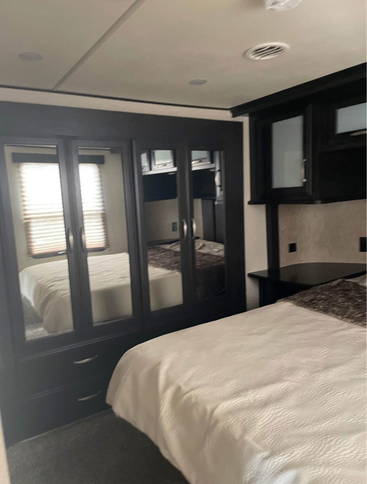 RV rental bedroom with white bed, dark wood furniture including mirrored wardrobe, and overhead cabinets for a cozy interior.. Grand Design Momentum Toy Hauler 2016