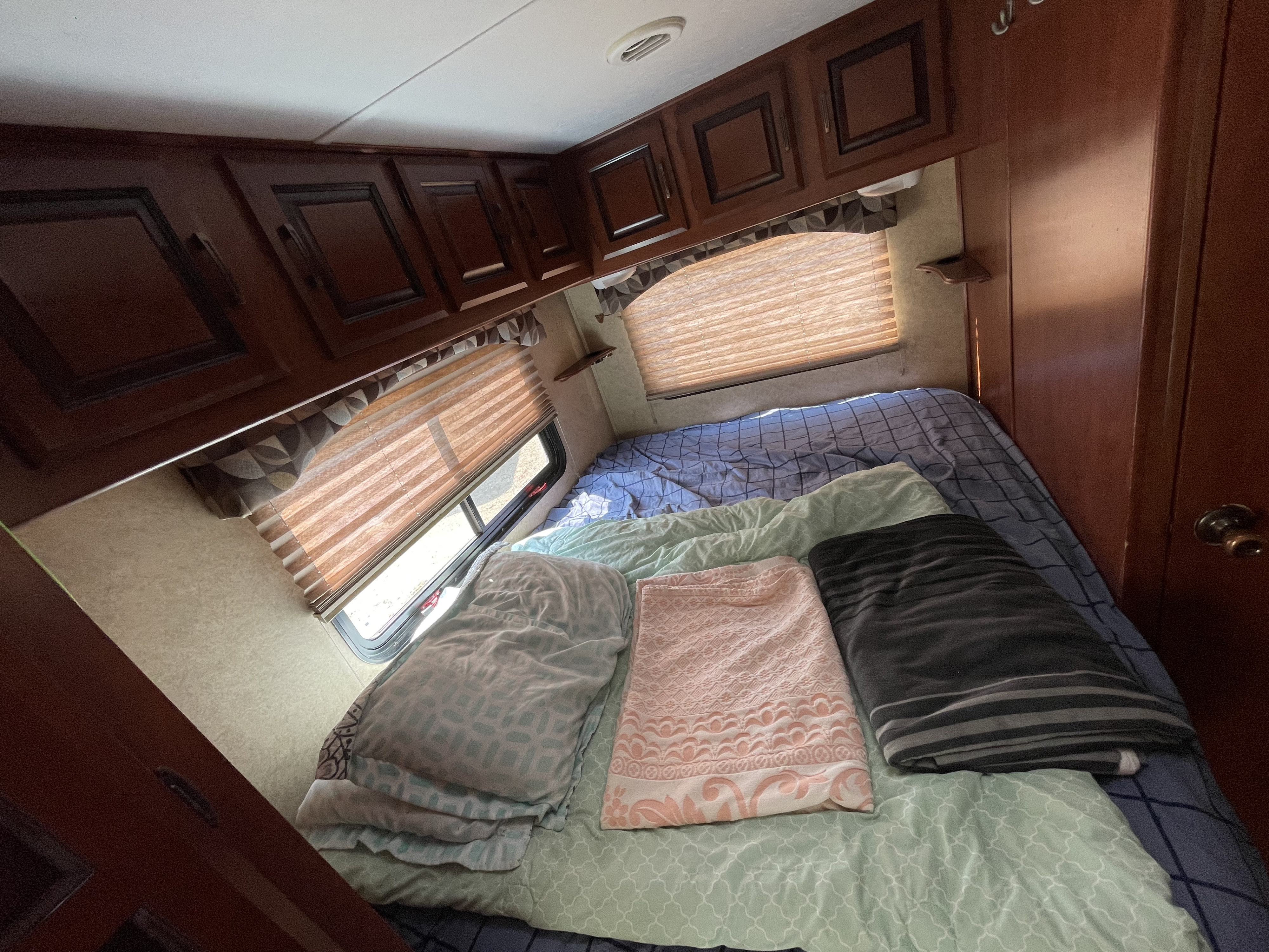 Cozy rv rental bedroom: bed with multiple blankets, window with blinds, and dark wood overhead cabinets.. Forest River Sunseeker 2011