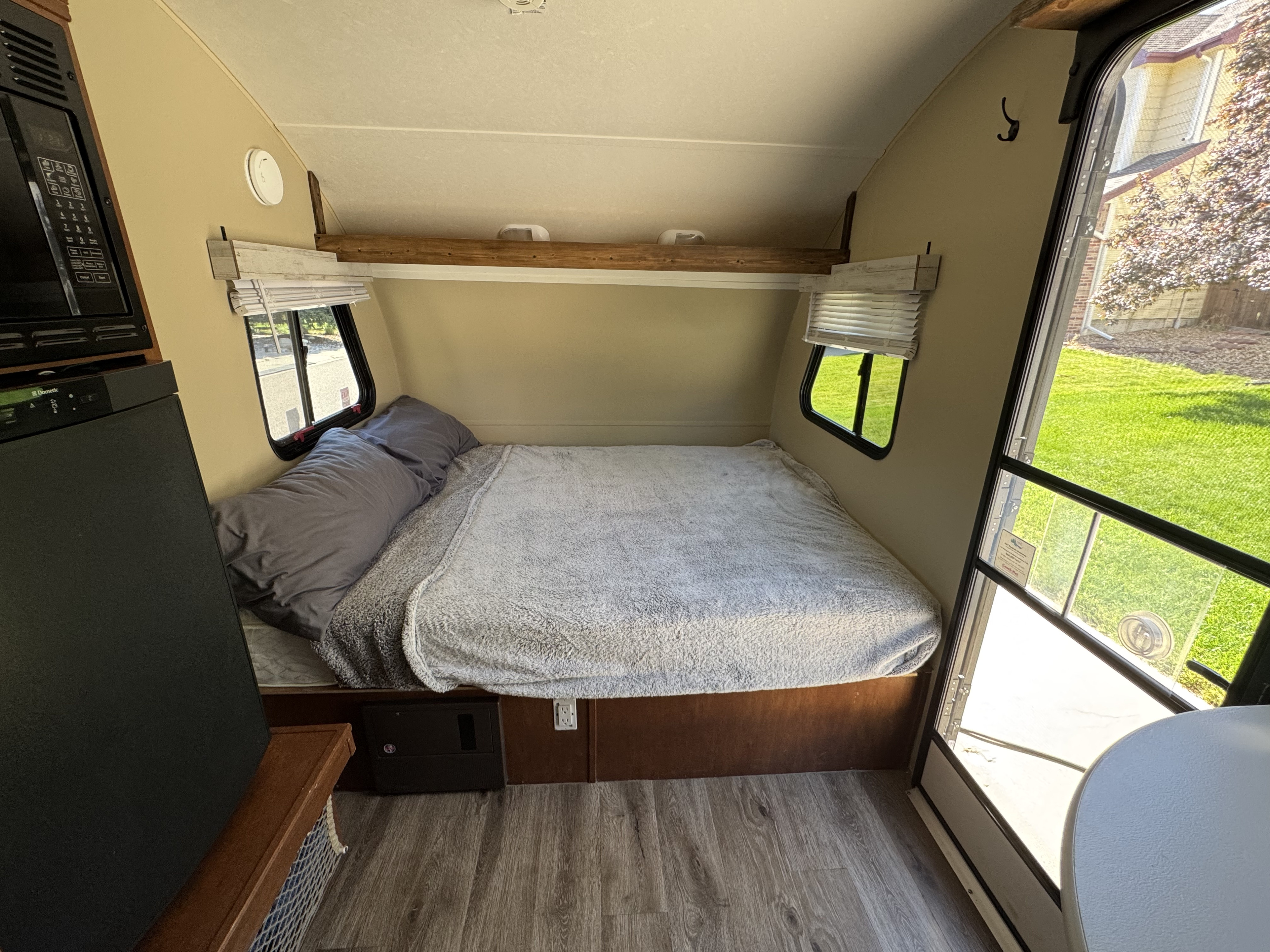 Cozy RV rental interior with a bed, microwave, Dometic fridge, overhead wooden shelf, and windows looking out to grass.. Forest River R-Pod 2013