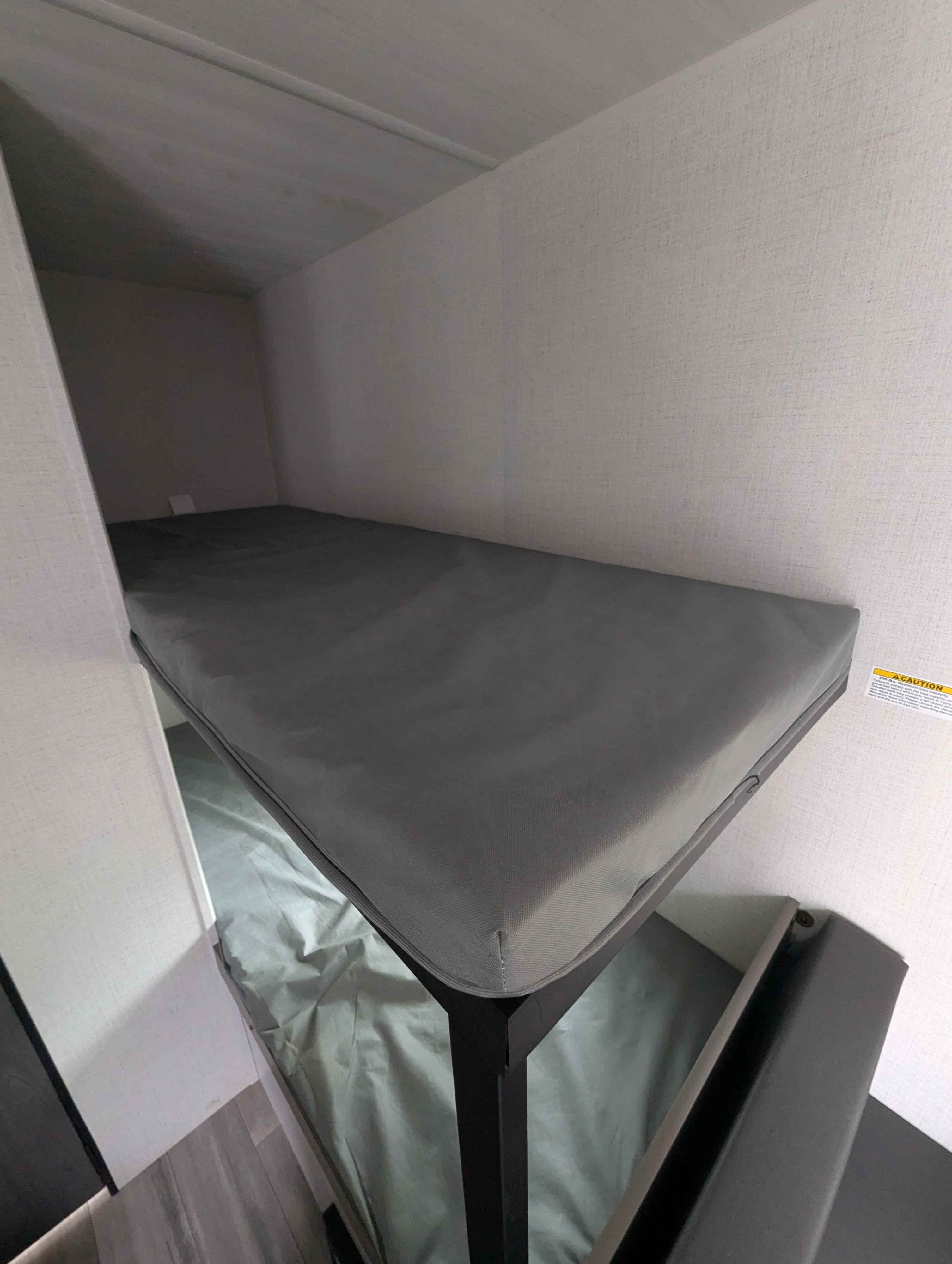 Inside an RV rental: a top bunk bed with grey mattress and lower bed, set against light textured walls.. Keystone Hideout 2024