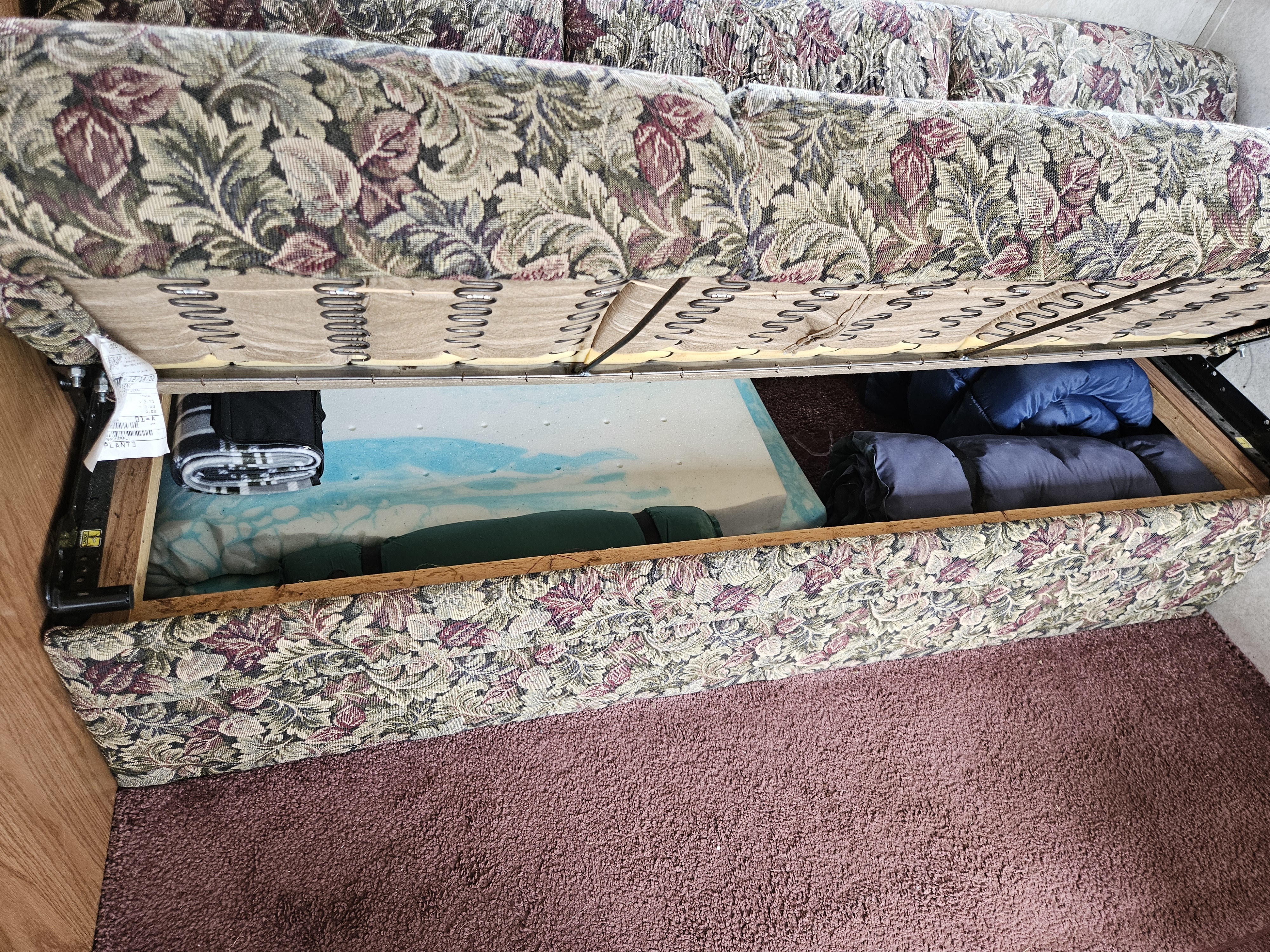 RV rental couch storage: Lifted floral cushion reveals hidden compartment with a mattress topper and rolled bedding.. Keystone RV Springdale 2002