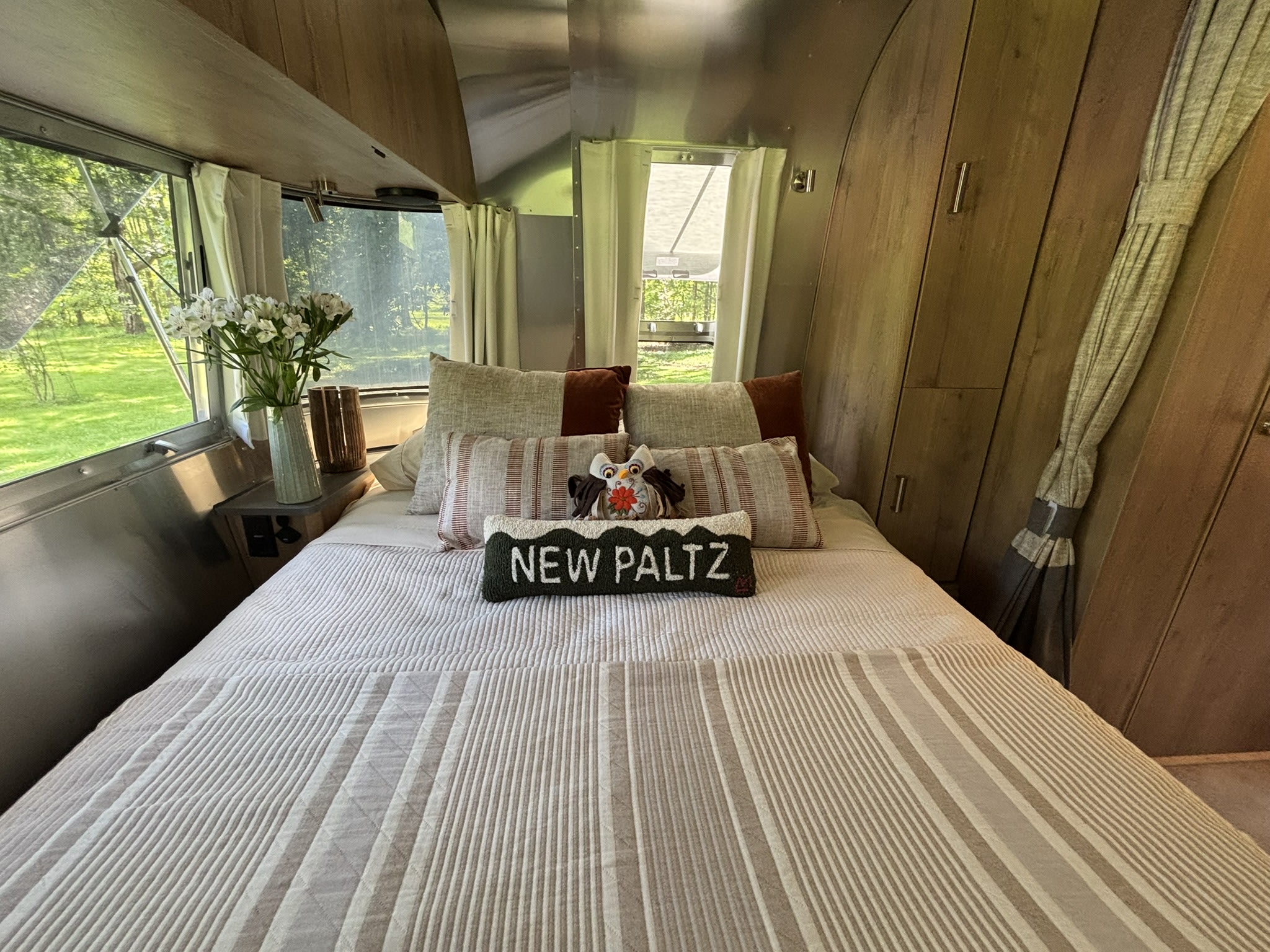 Airstream RV rental bedroom: striped bedding, "NEW PALTZ" pillow, and lush outdoor views. A cozy travel retreat.. Airstream Tradewinds 2025
