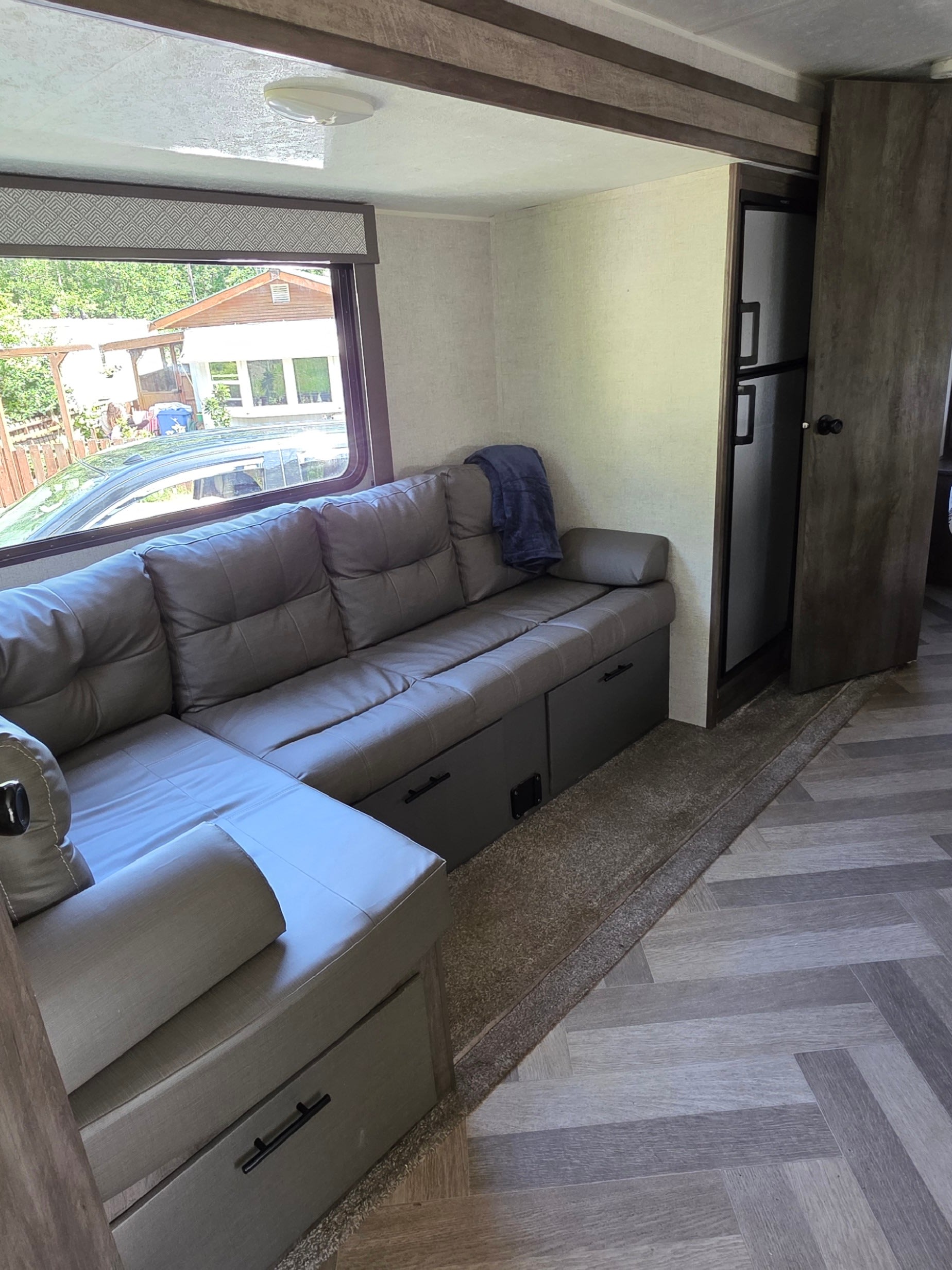 Modern RV rental living area featuring a large grey L-shaped sofa with storage, a window view, and fridge.. Forest River Wildwood 2020