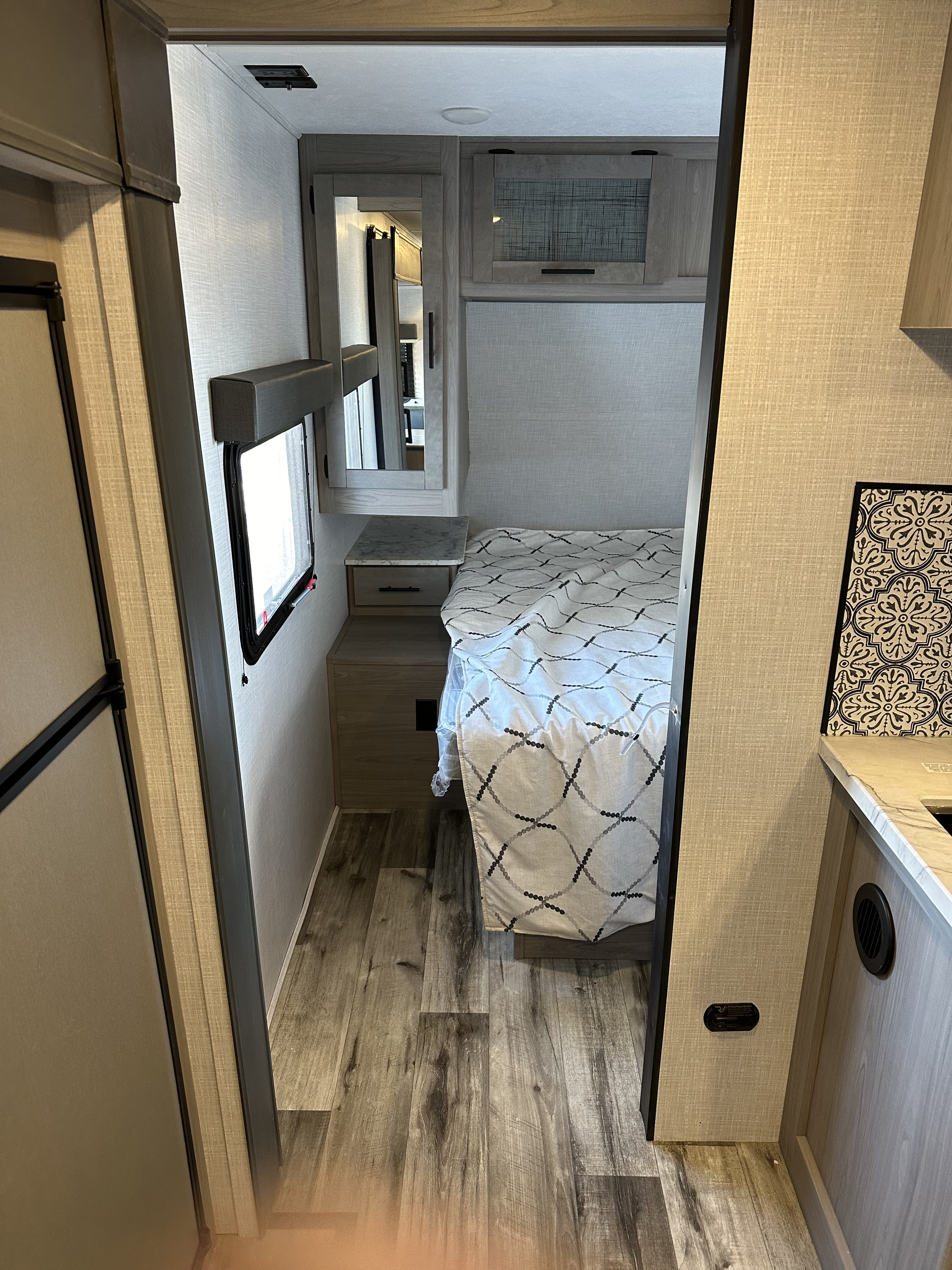View into a modern RV rental bedroom, featuring a patterned bedspread, nightstand, overhead cabinet, and wood-look floor.. Coleman Dutchman Light 2023