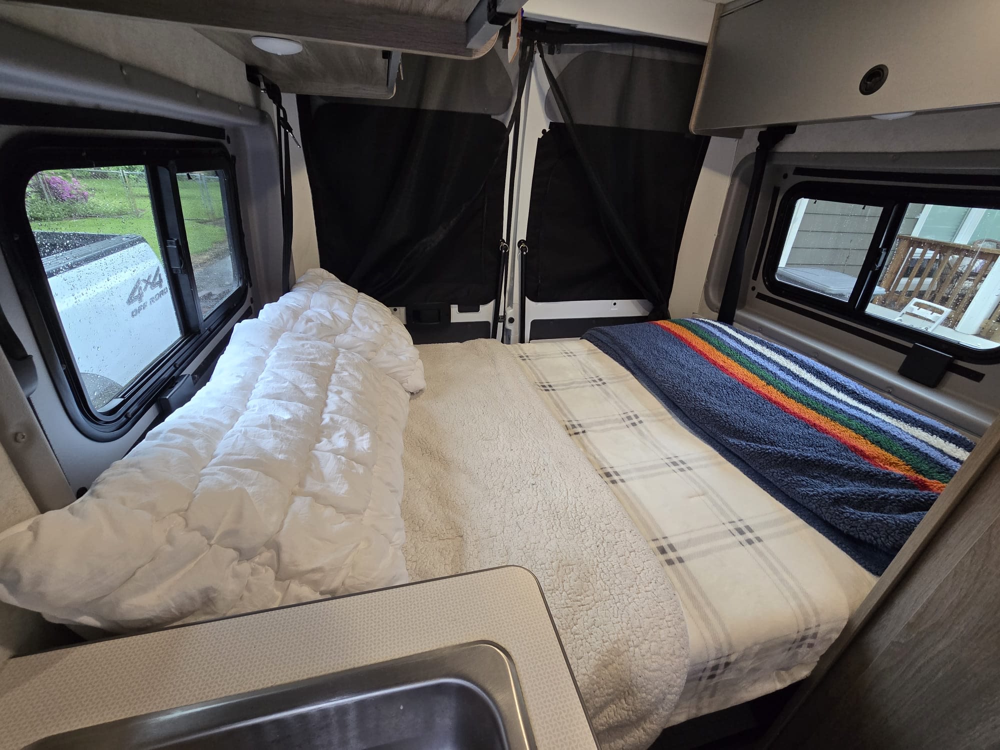 Cozy RV rental interior featuring a comfy bed with plaid and rainbow blankets, a sink, and window views.. Winnebago Solis 2023