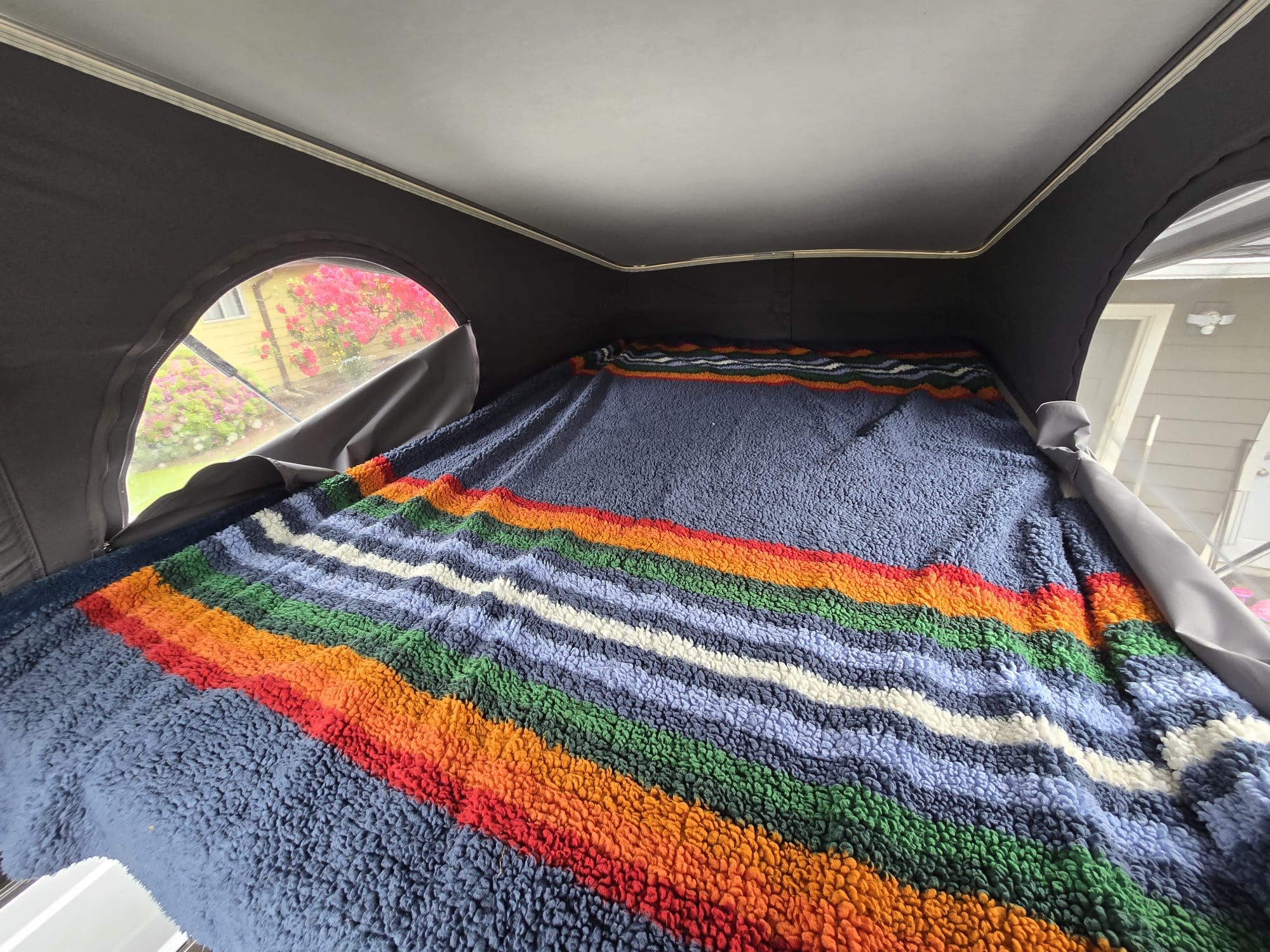 Inside a cozy RV rental pop-top, a colorful textured blanket covers the bed, with a view of blooming flowers.. Winnebago Solis 2023