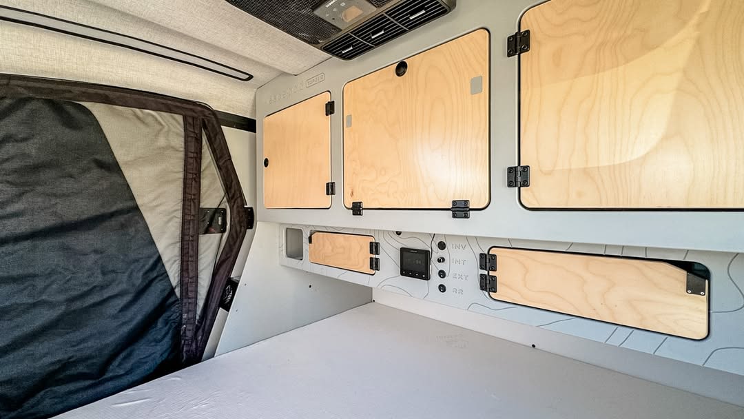 Modern rv rental interior: organized with light wood overhead storage, a system control panel, and a cozy bed area.. Escapod Topo 2 2024