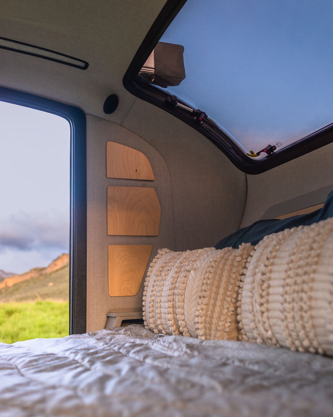 Inside a cozy RV rental: a comfortable bed, wooden shelves, and a large skylight showing blue sky, with a scenic outdoor view.. Escapod Topo 2 2024