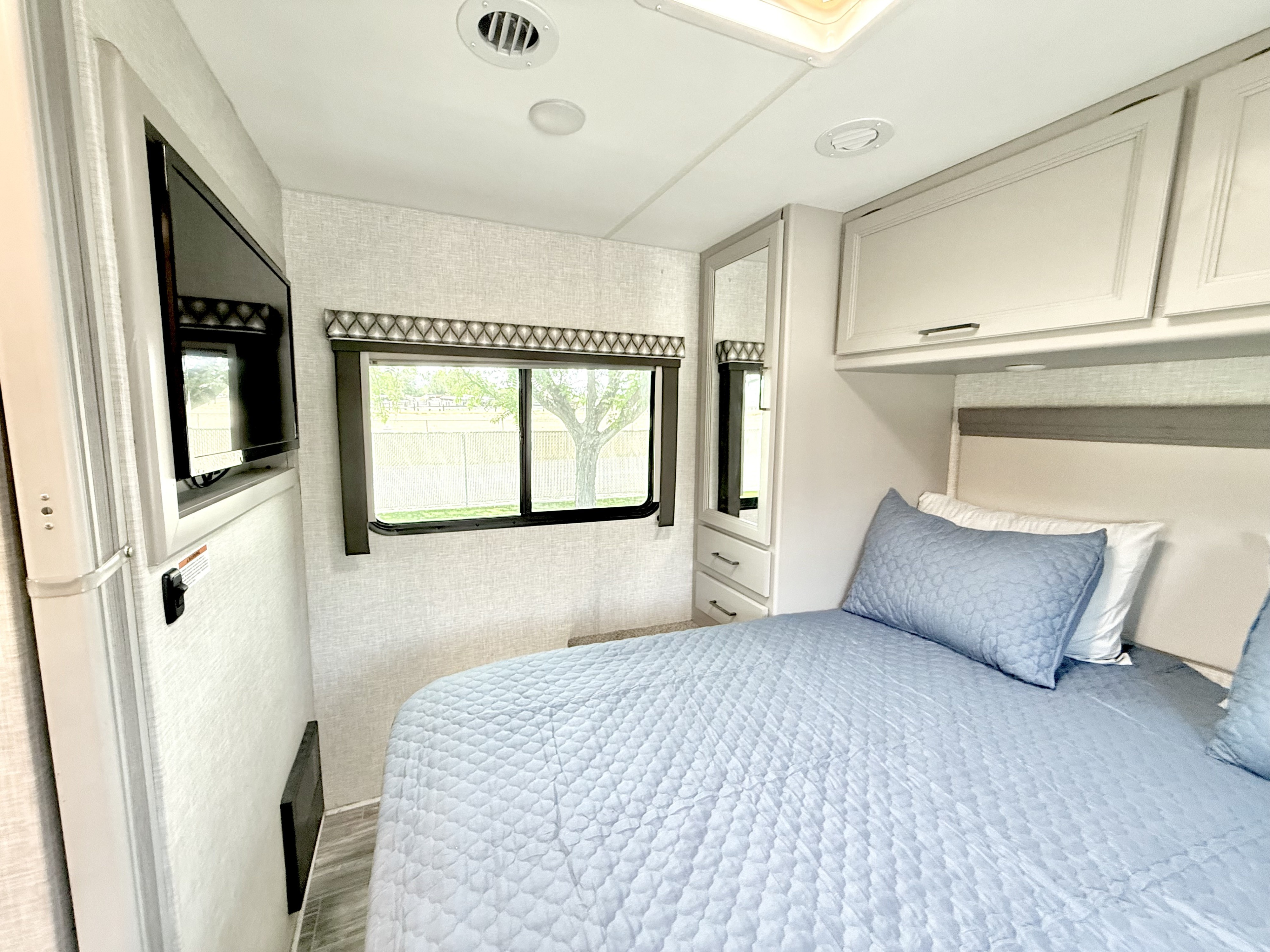 Bright RV rental bedroom featuring a bed with blue quilted bedding, window, TV, and built-in storage.. Thor Geneva 2022