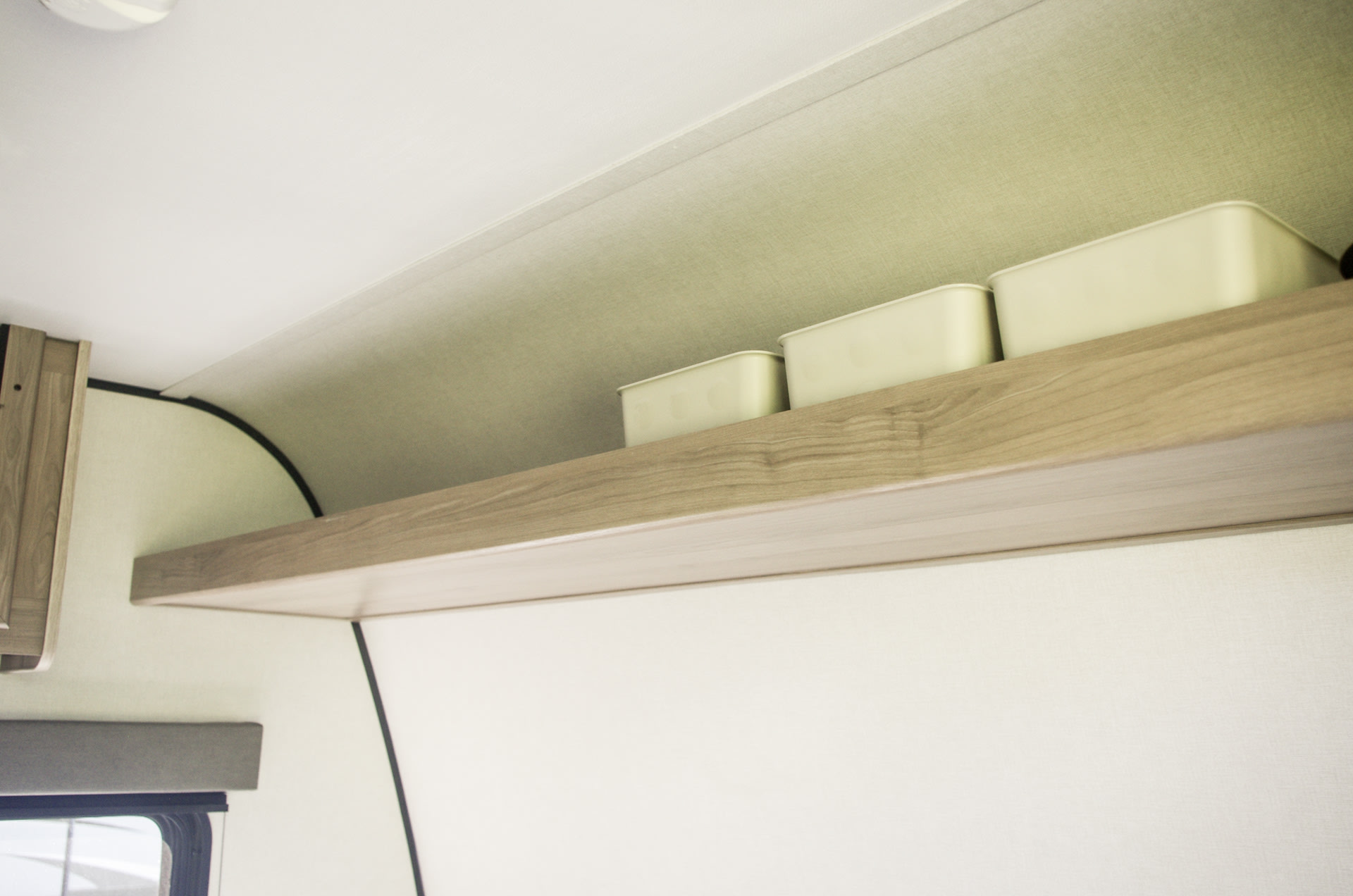 RV rental cabin interior features a light wood shelf with three pale storage bins. Above are curved light-colored panels.. Keystone Coleman 13B 2025