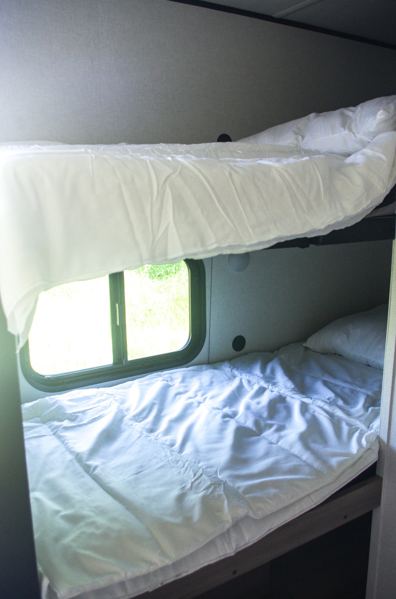 Clean, cozy bunk beds with white linens in an RV rental. A window reveals green nature outside, ideal for travel.. Keystone Coleman 13B 2025