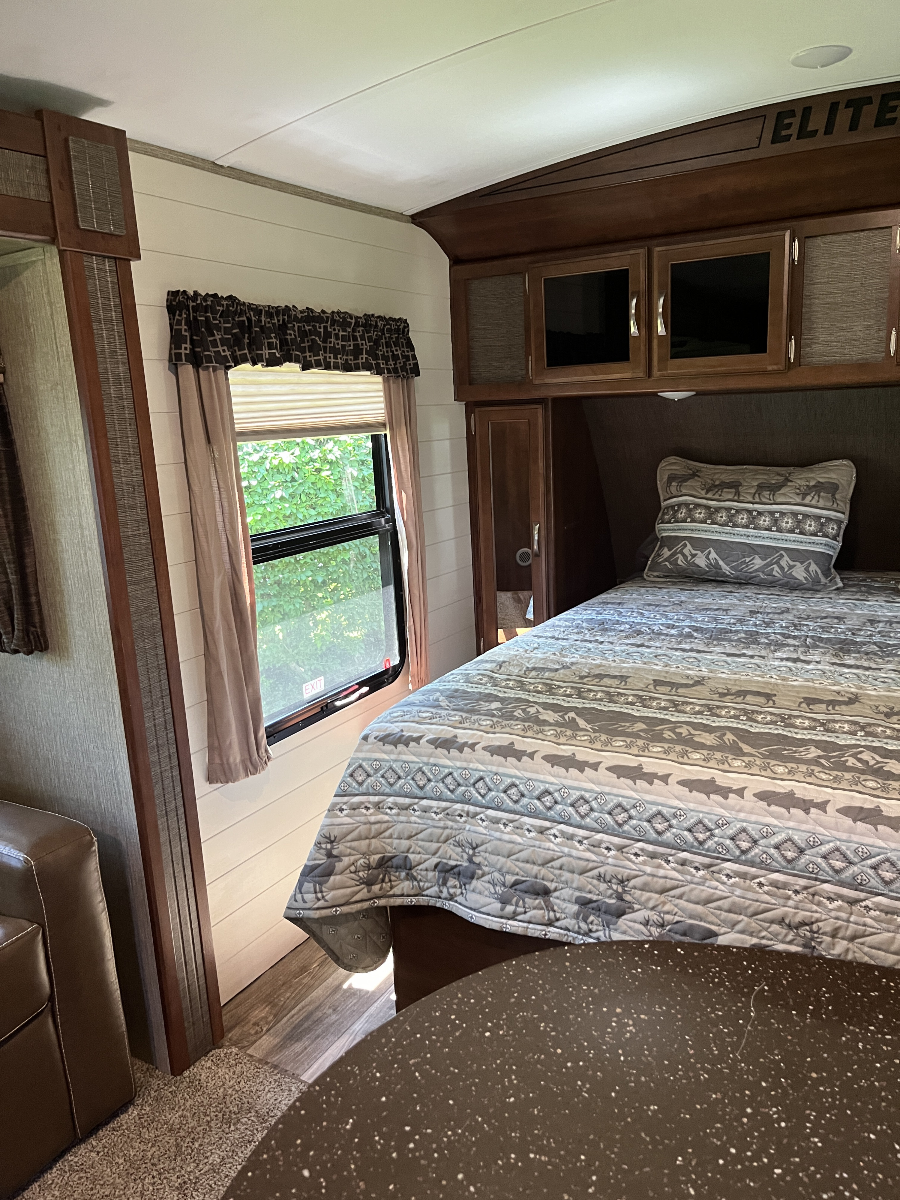Cozy RV rental bedroom with nature-themed bedding, overhead storage, and a bright window looking out to greenery.. Keystone RV Passport Elite 2017