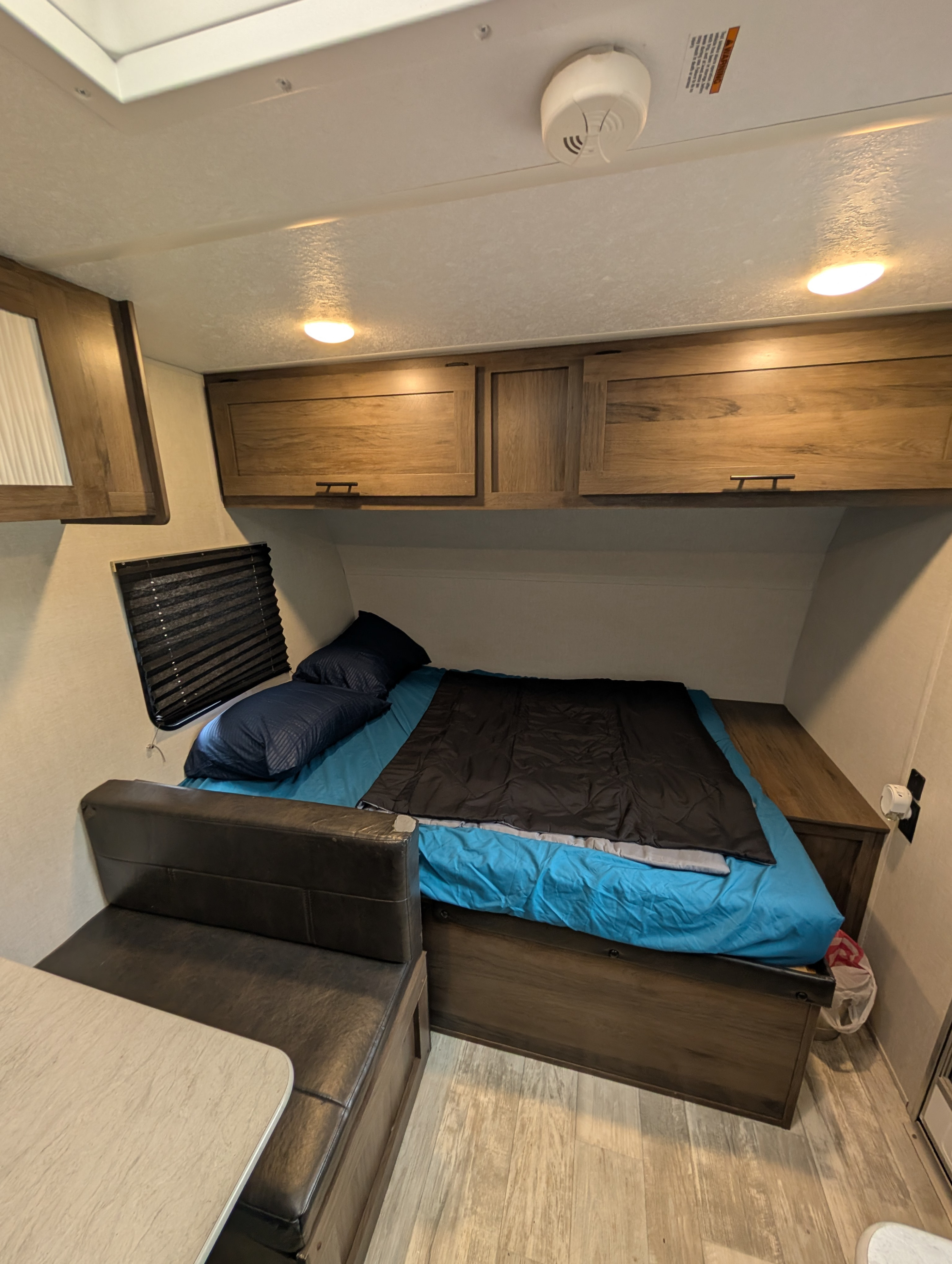 Inside an RV rental: a compact living space featuring a neatly made bed with blue bedding, overhead wooden cabinets, and a small dinette area.. Avenger LT 2021