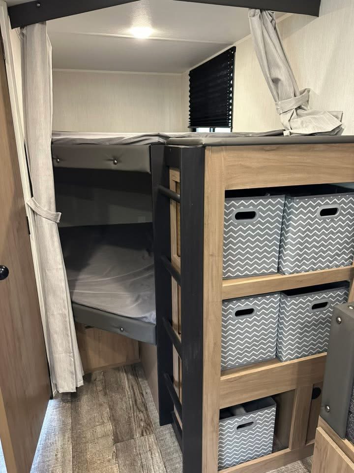RV rental interior: cozy bunk beds with privacy curtains, a ladder, storage bins, and wood-look flooring.. Forest River Campsite Ultra 26CJL 2025