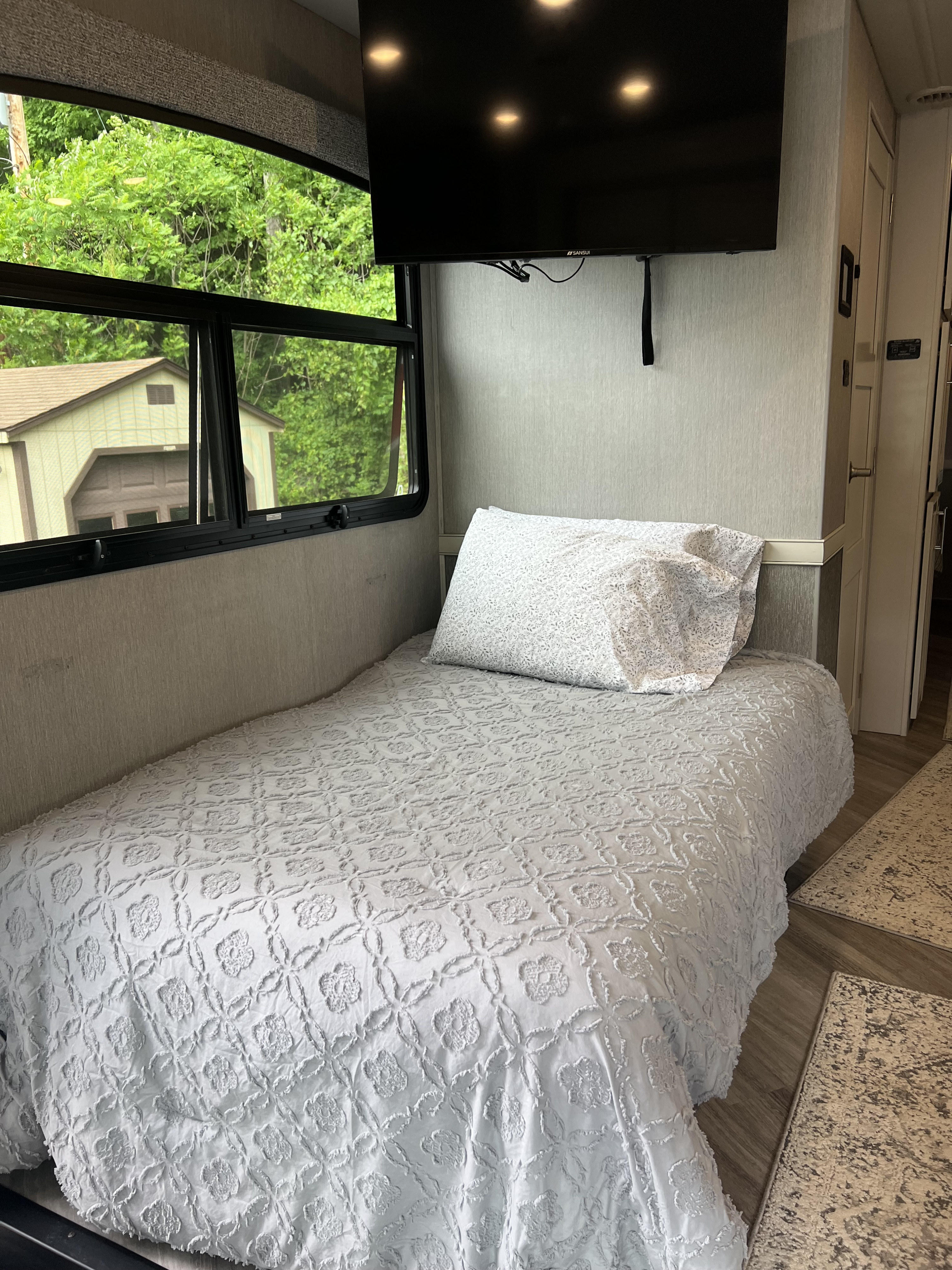 Comfortable RV rental interior with a cozy bed, large window showcasing lush greenery, and a mounted TV. Perfect for travel!. Fleetwood Fortis 2024