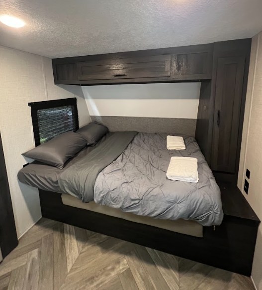 Cozy RV rental bedroom featuring a queen bed with grey bedding, overhead storage, and a small window for natural light.. Forest River ozark 2021