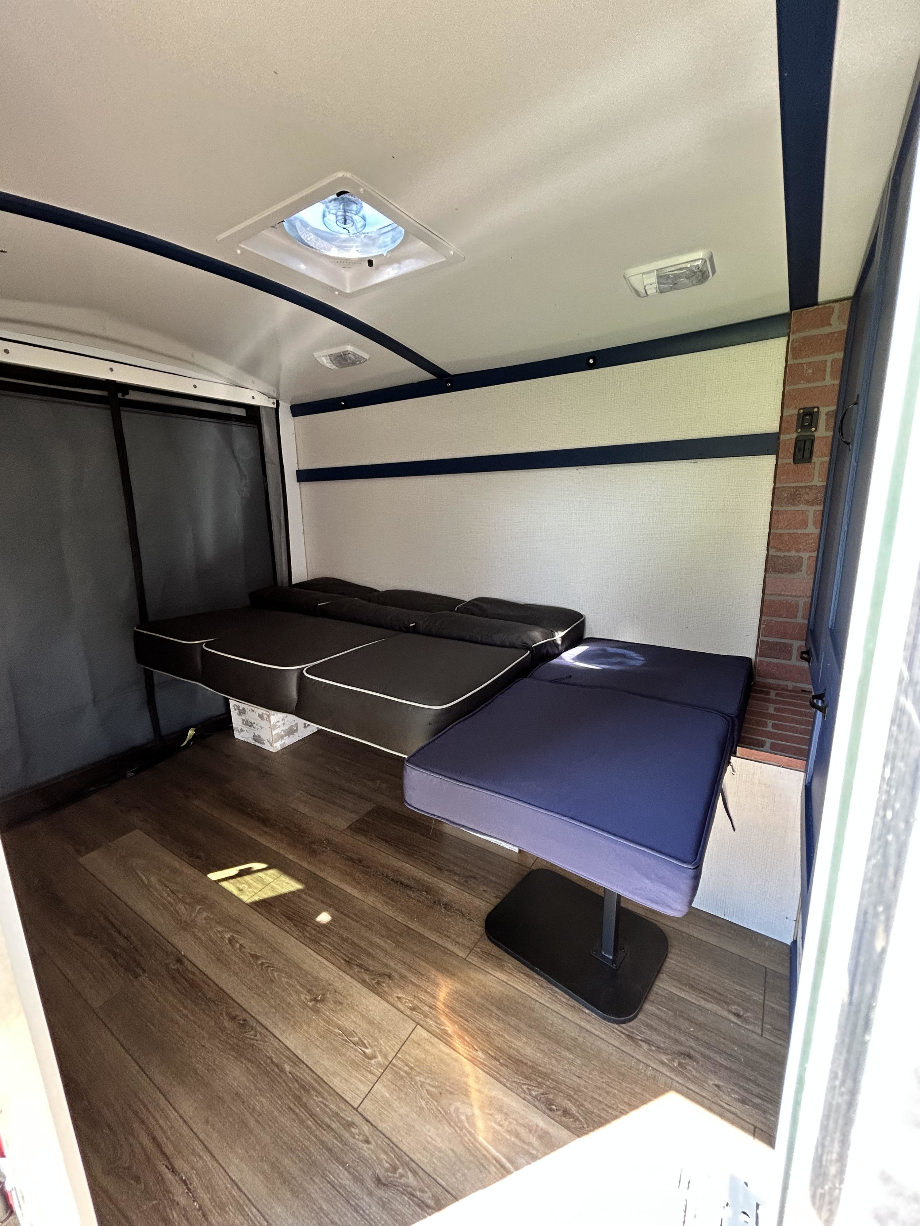 Inside an RV rental, showing convertible black and navy seating/beds on wood laminate flooring. Features a ceiling fan and interior lights.. Custom Camper 1999