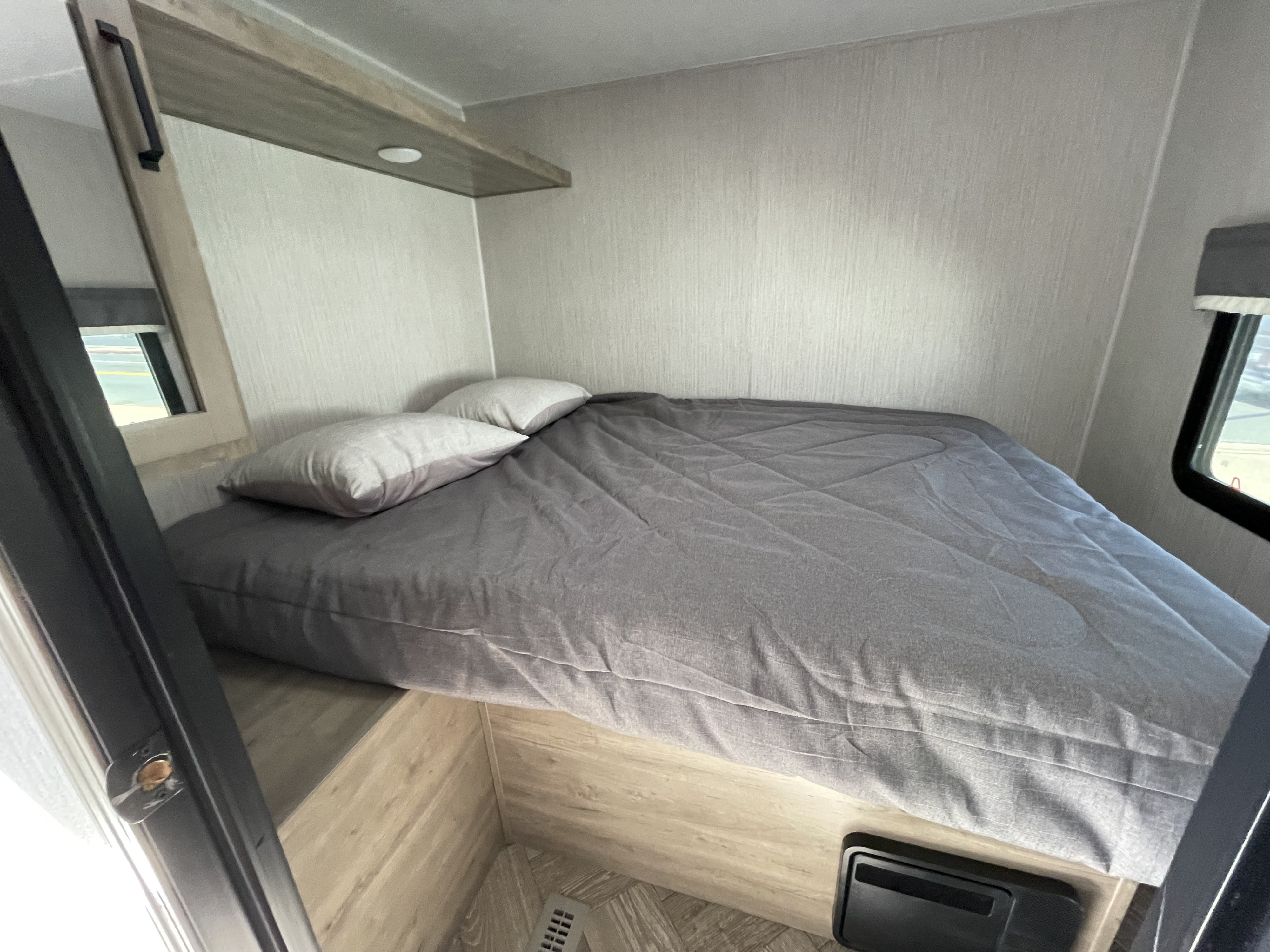 Inside an RV rental bedroom: a comfortable bed with grey bedding, a wall-mounted shelf, and a small window.. Forest River Palomino Puma 2021
