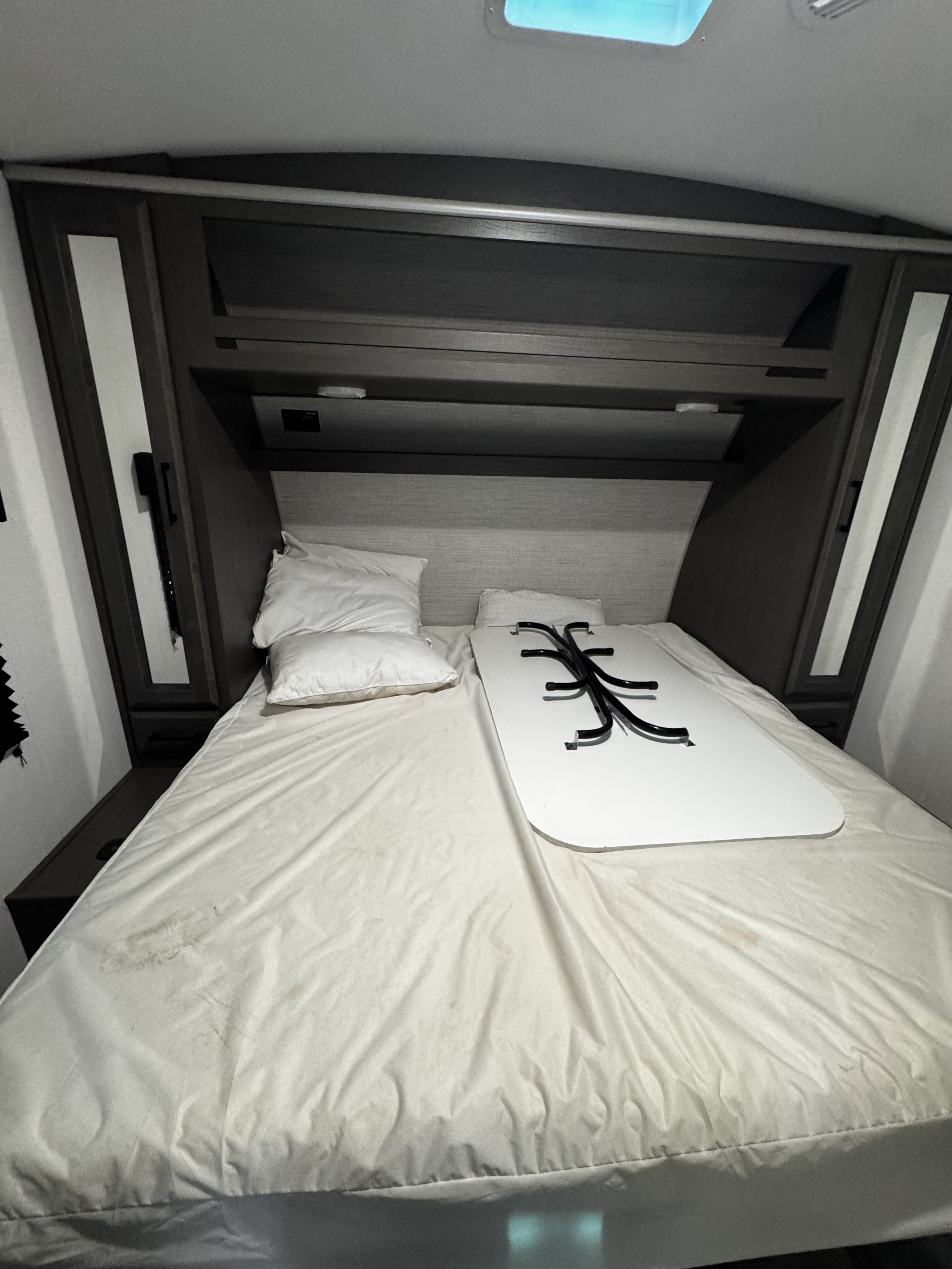 RV rental bedroom: white bed with pillows, dark wood cabinets, and a portable table stored on the bed. Cozy interior.. Heartland Fuel 287 2021