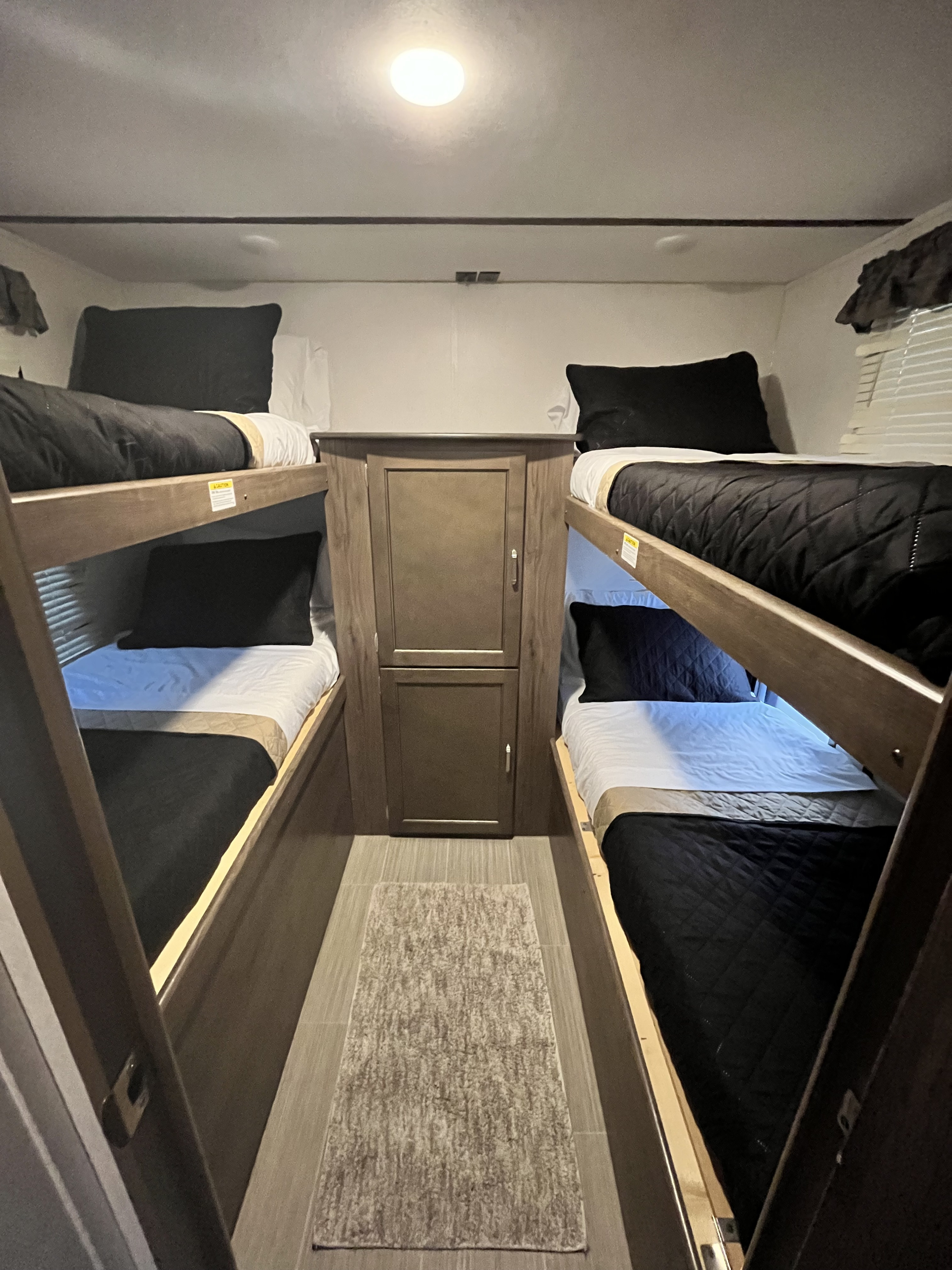 Inside an RV rental, this bunk room features two sets of neatly made bunk beds with black and white linens, separated by a central storage unit, with a small rug on the floor.. Keystone RV Passport Grand Touring 2019