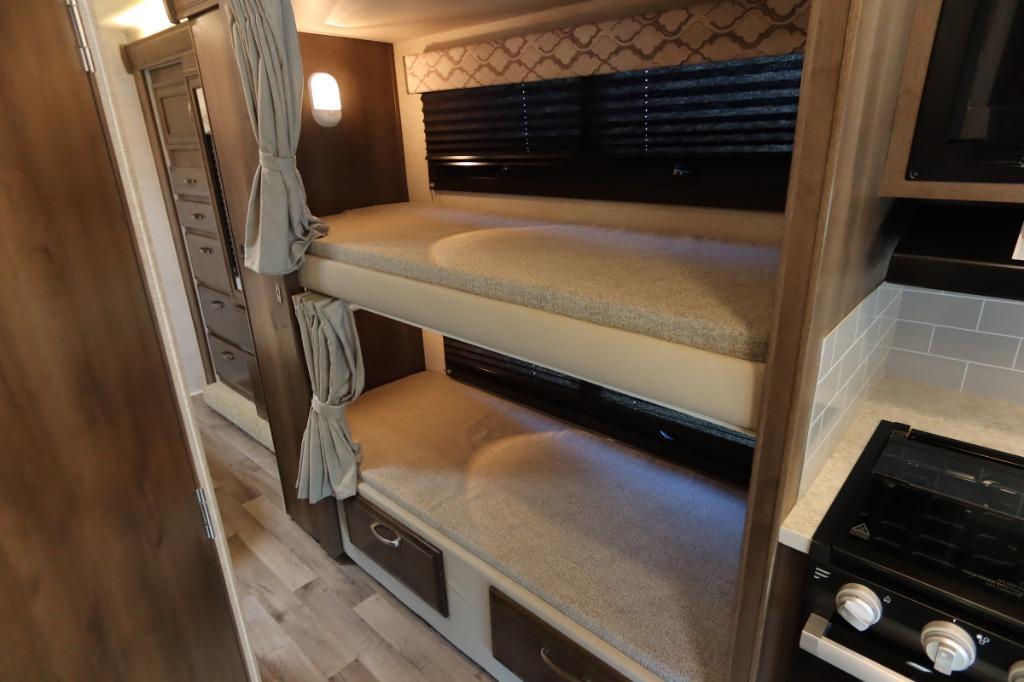 This RV rental features two stacked bunk beds with privacy curtains, adjacent storage, and a compact kitchen with a stove.. Jayco Redhawk 2021