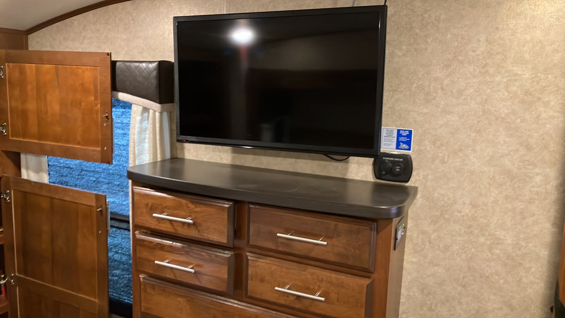 RV rental bedroom featuring a wall-mounted TV, wooden dresser, and open cabinets revealing a patterned shade.. Jayco Eagle 2018