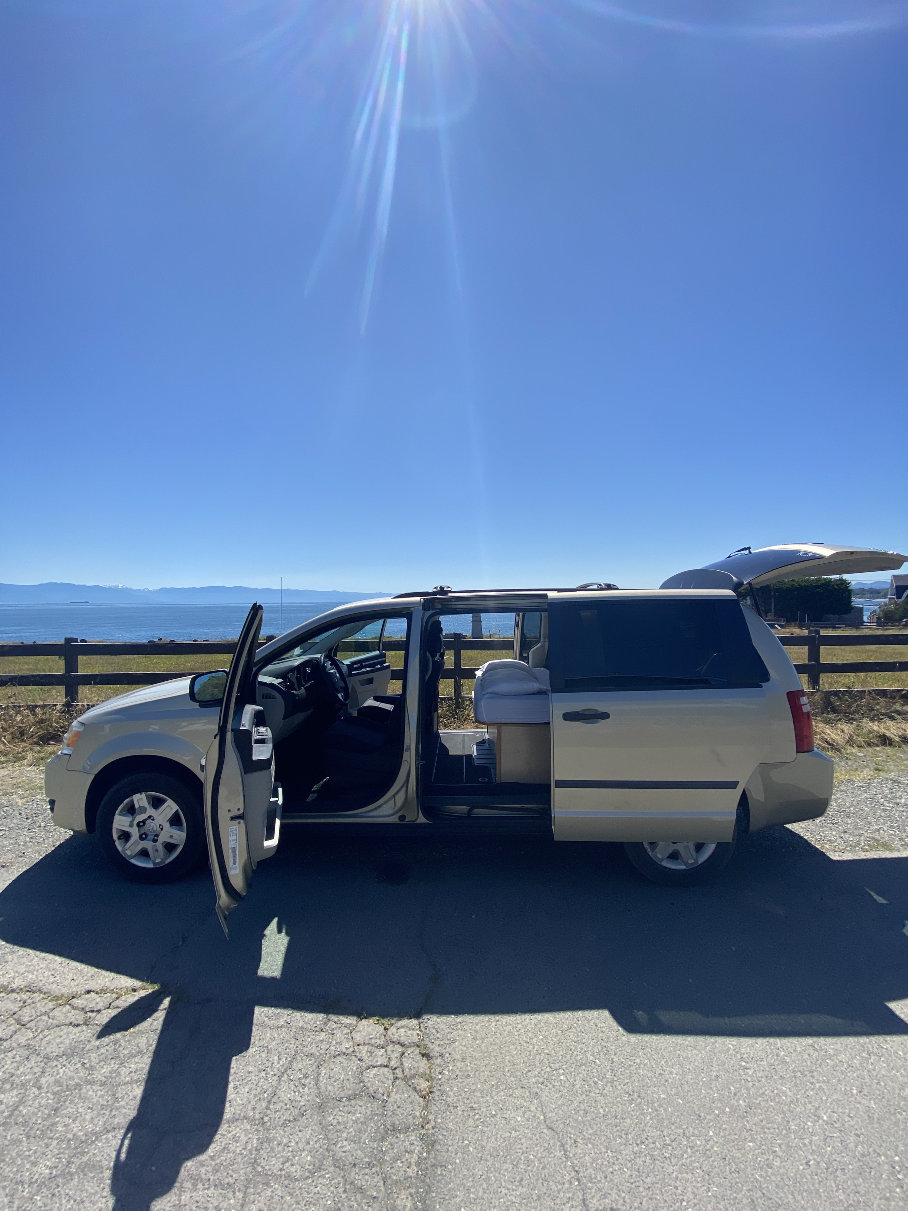 Minivan camper RV rental with doors open, showing bed setup. Parked by ocean, ready for road trips.. Dodge Grand Caravan SE 2010