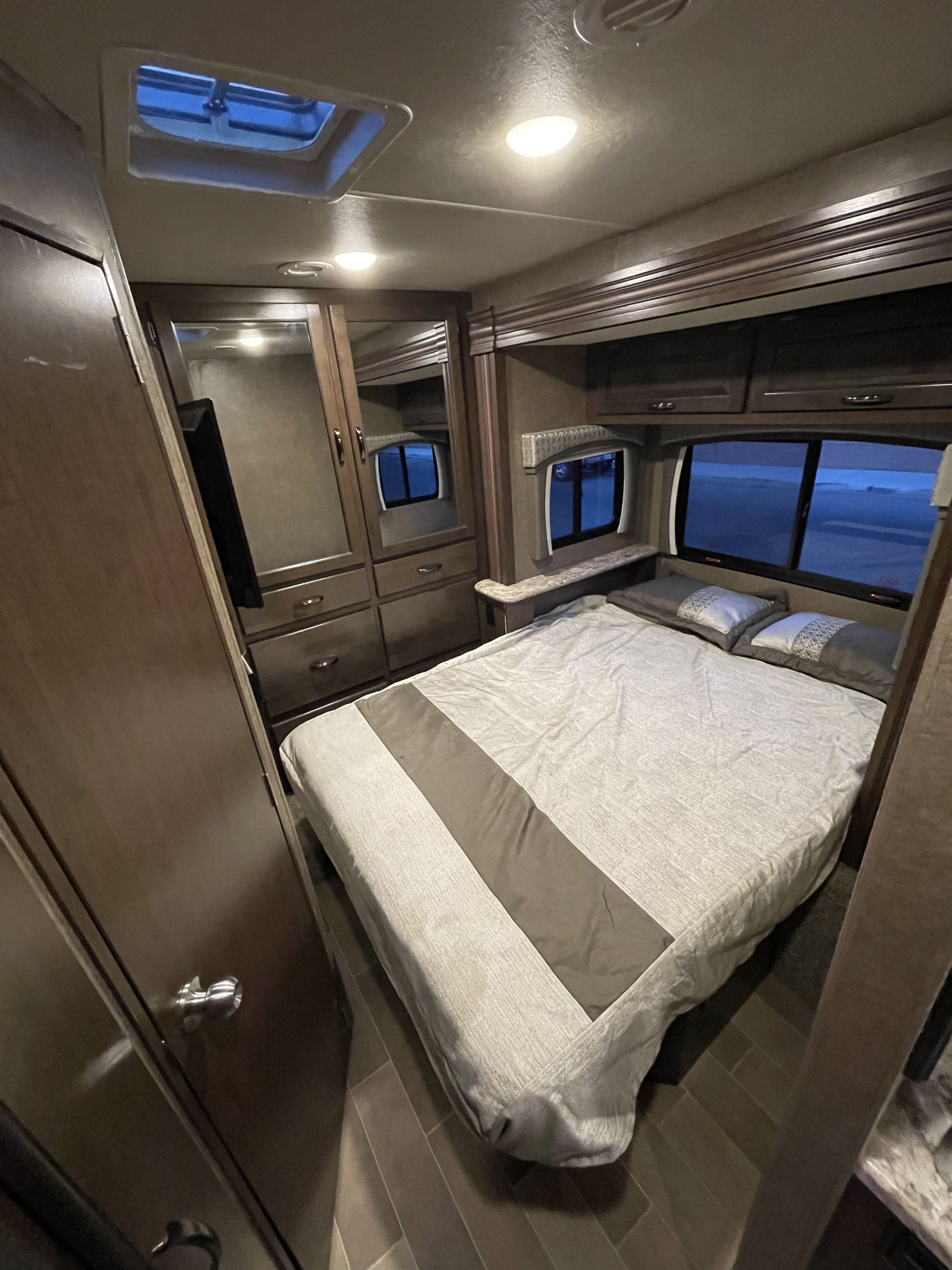 RV rental bedroom interior with a neatly made bed, dark wood cabinets, and windows revealing the outside.. Thor Chateau Sprinter 2020