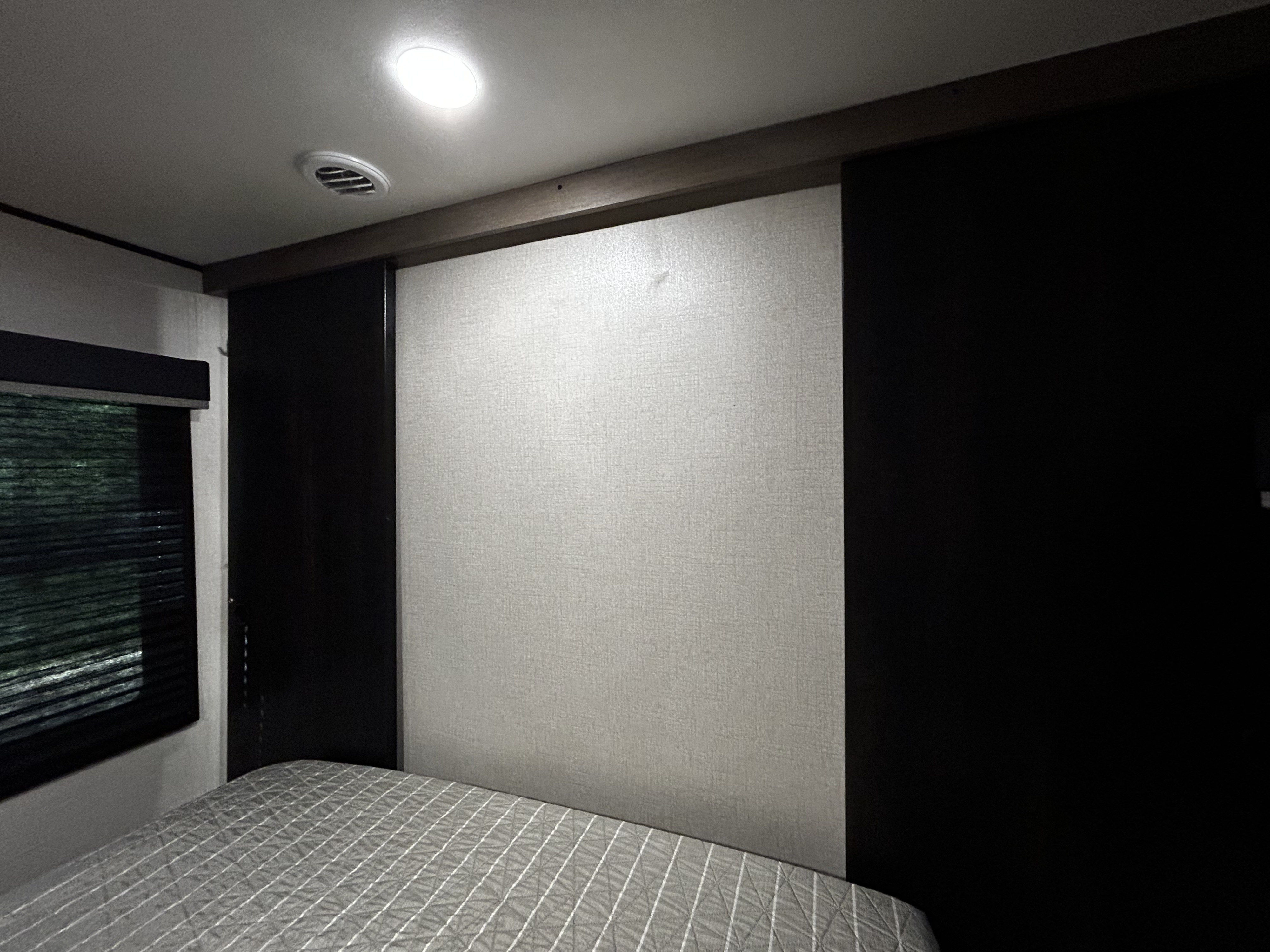 Compact RV rental bedroom featuring a bed with a grey comforter, light headboard, window with blinds, and ceiling fixtures.. Highland Ridge RV BHS26 2023
