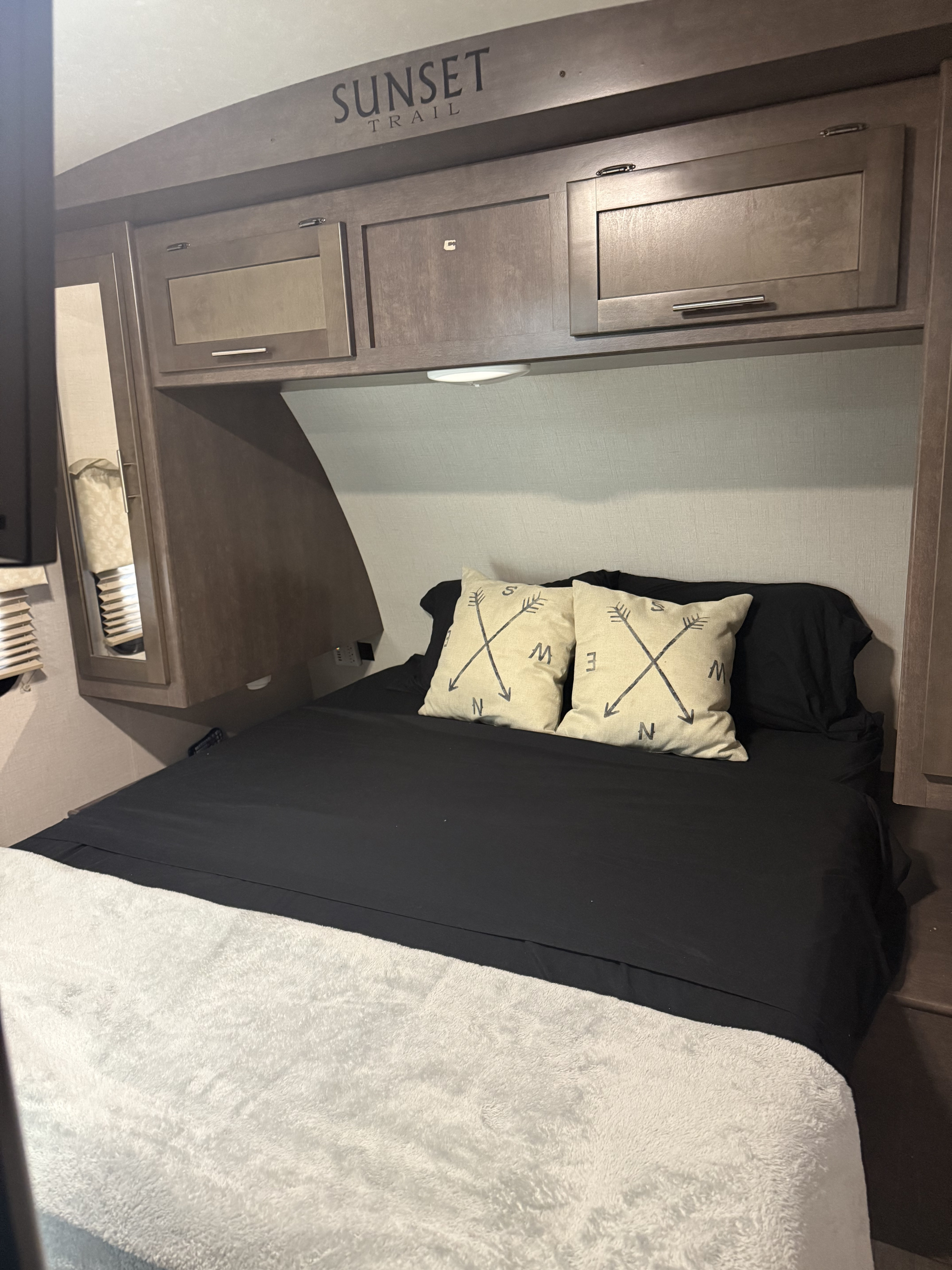 Inside an RV rental: a neat bedroom with a dark bed, fluffy blanket, arrow pillows, and wood cabinets. "Sunset Trail" branding.. Crossroads RV Sunset Trail Super Lite 2018