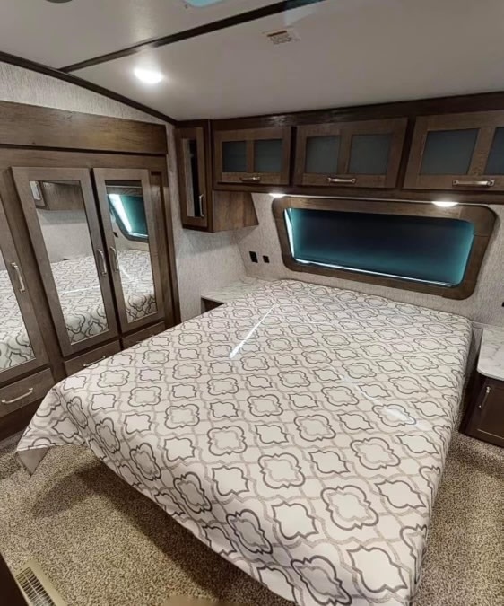 Modern RV rental bedroom featuring a patterned bed, mirrored wardrobe, and overhead cabinets with a large window.. Dutchmen Astoria 2021