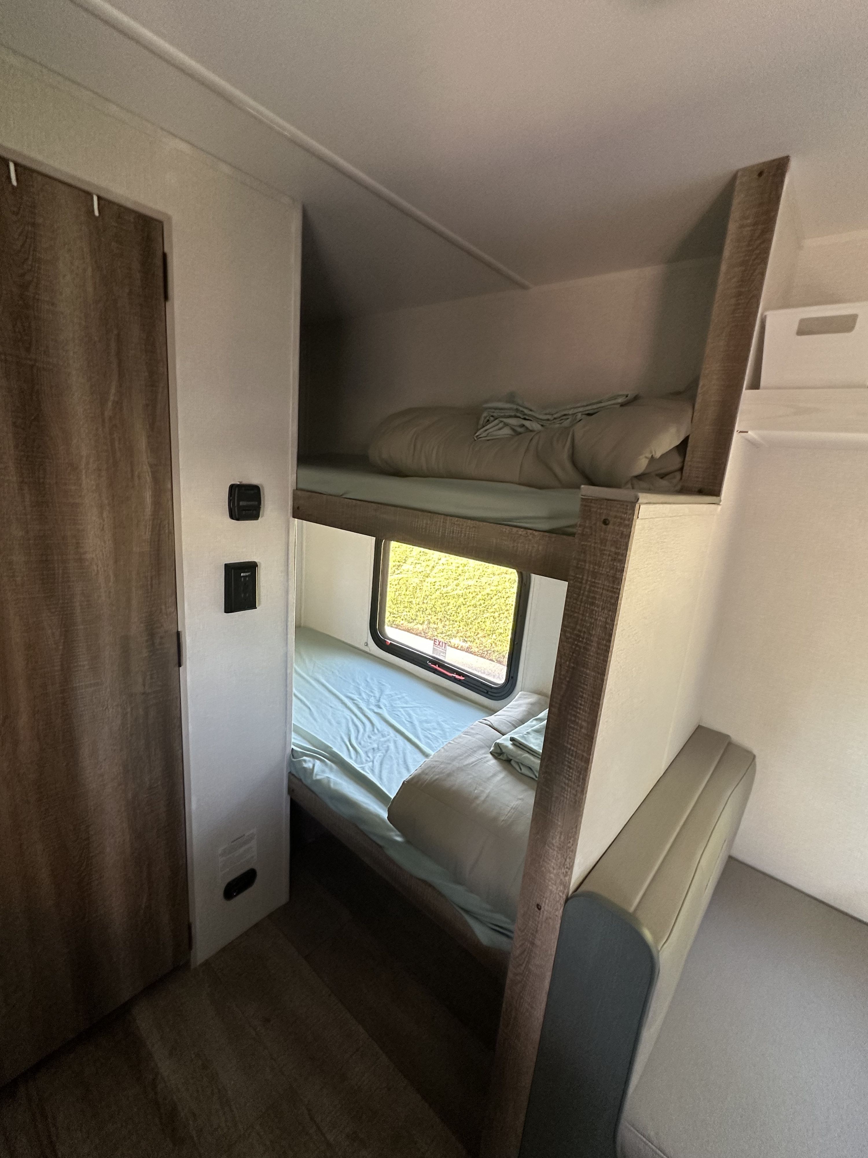 This RV rental interior features two bunk beds with light bedding, a window overlooking greenery, and a wooden door. Compact, functional sleeping.. Heartland Eddie Bauer 2025
