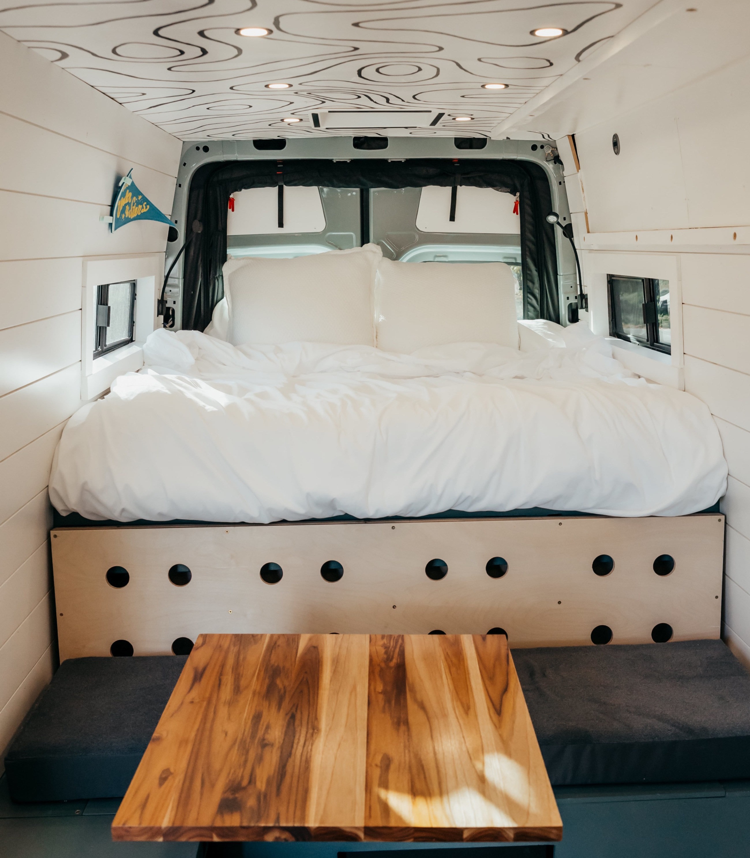 Inside a modern RV rental: a large, neatly made bed dominates, with a wooden table & upholstered bench in the foreground. Topographic ceiling.. Mercedes-Benz Sprinter 2022