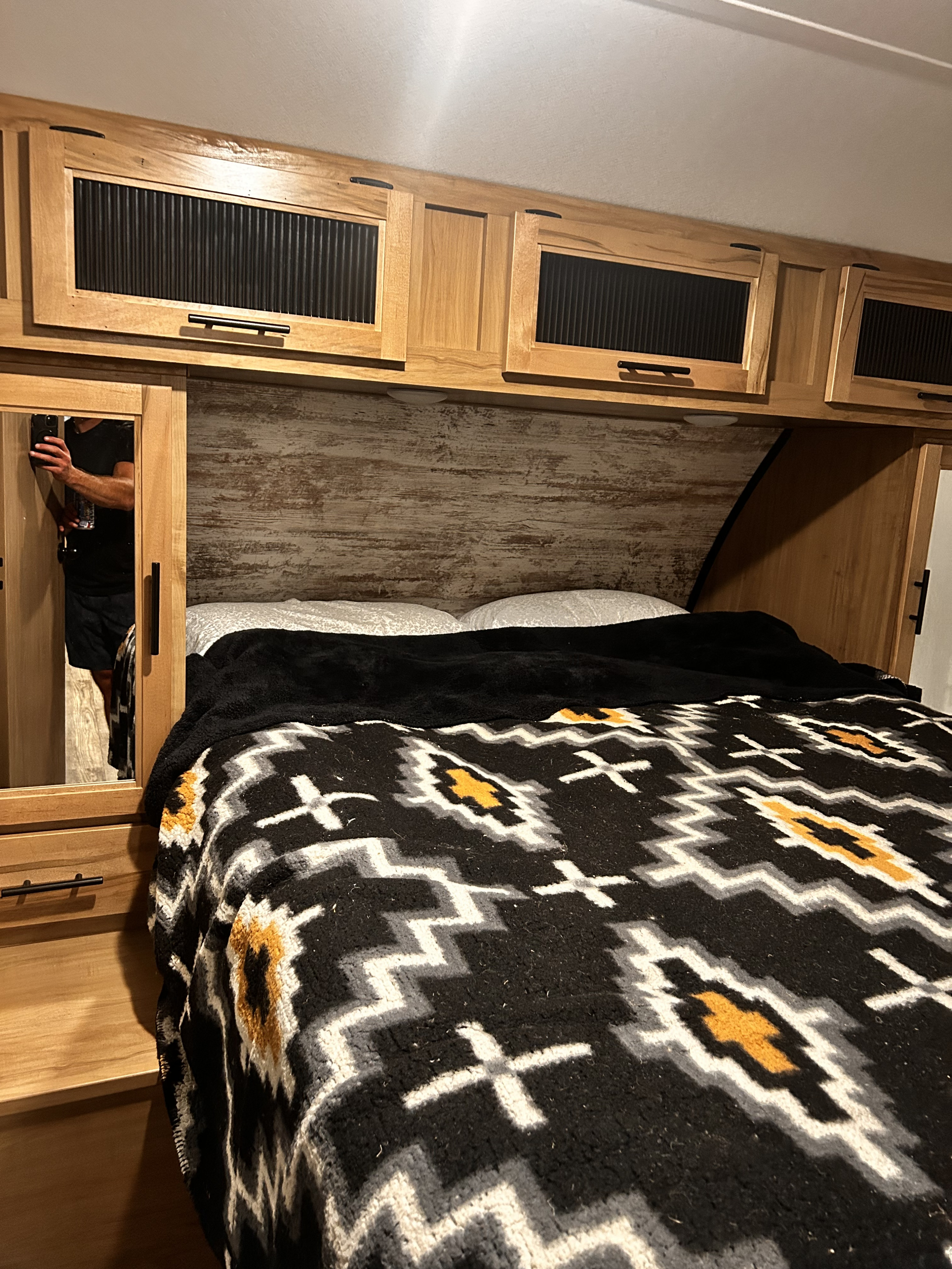 Cozy RV rental bedroom: light wood cabinets, distressed headboard, black/gold patterned blanket, and mirrored wardrobe.. Forest River Cherokee Alpha Wolf DBH-L 2024