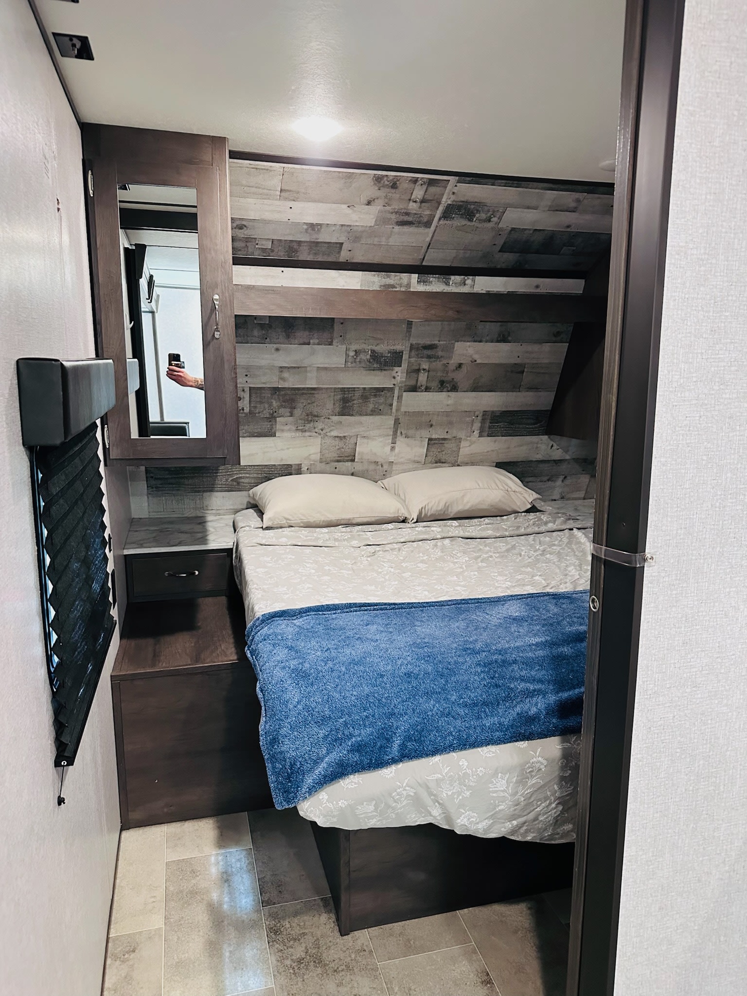 Cozy rv rental bedroom with a bed, blue blanket, wood-paneled wall, nightstand, mirror, and tiled floor.. Dutchmen Coleman Lantern 2021