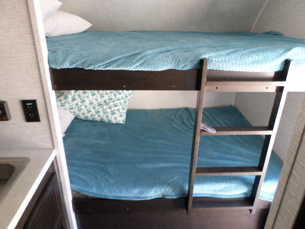 This RV rental features bright bunk beds with blue blankets and a ladder. A compact sink area is visible on the left for comfortable travel.. Braxton Creek Bushwhacker Plus 2022