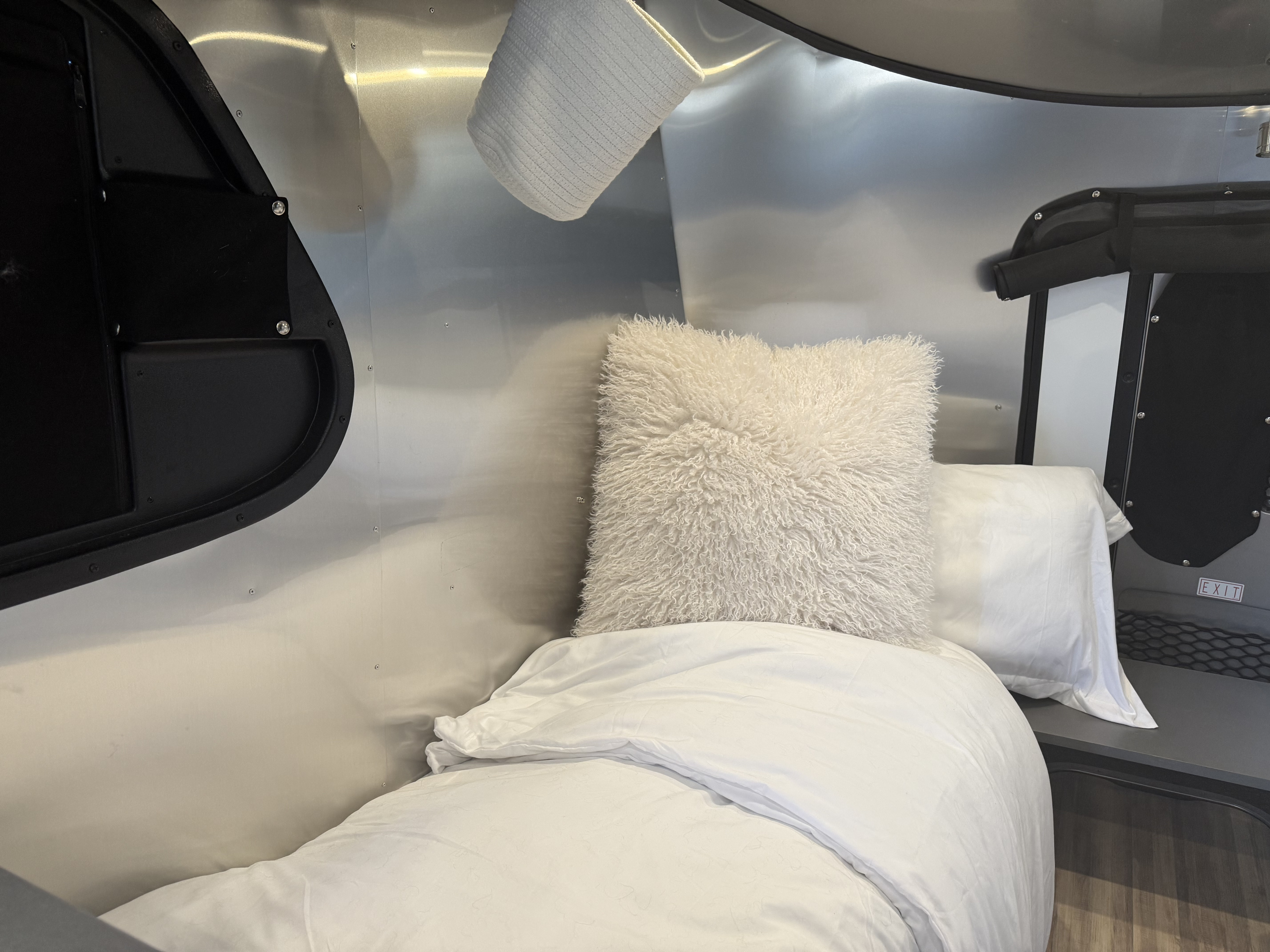 RV rental interior: a comfy bed with white linens & fluffy pillow, set against sleek metal walls & modern black panels.. Airstream Basecamp 2023