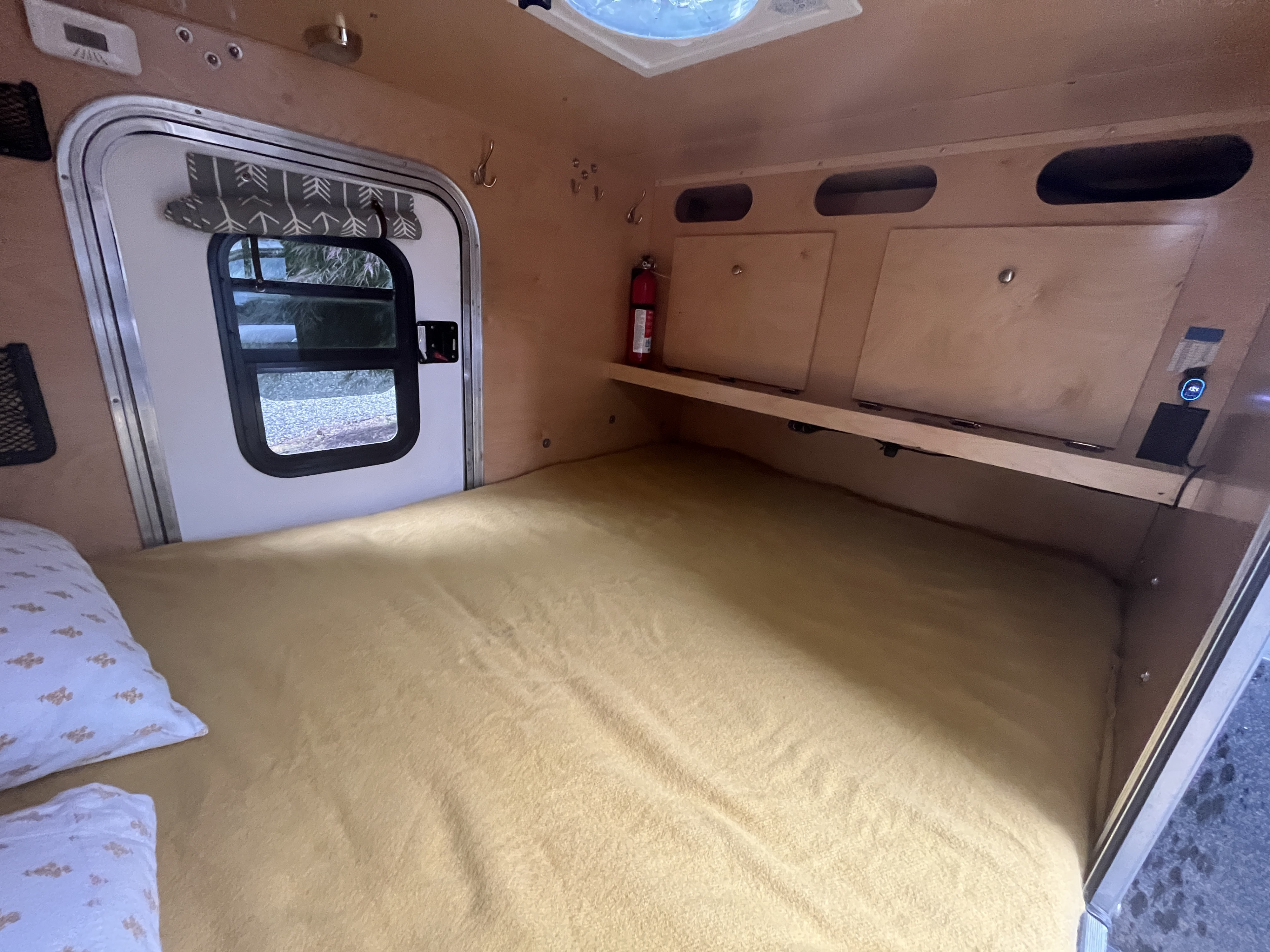 Interior view of a compact rv rental camper showing a cozy bed, side door with window, overhead storage, and a fire extinguisher.. Teardrops NW Ranger 2019