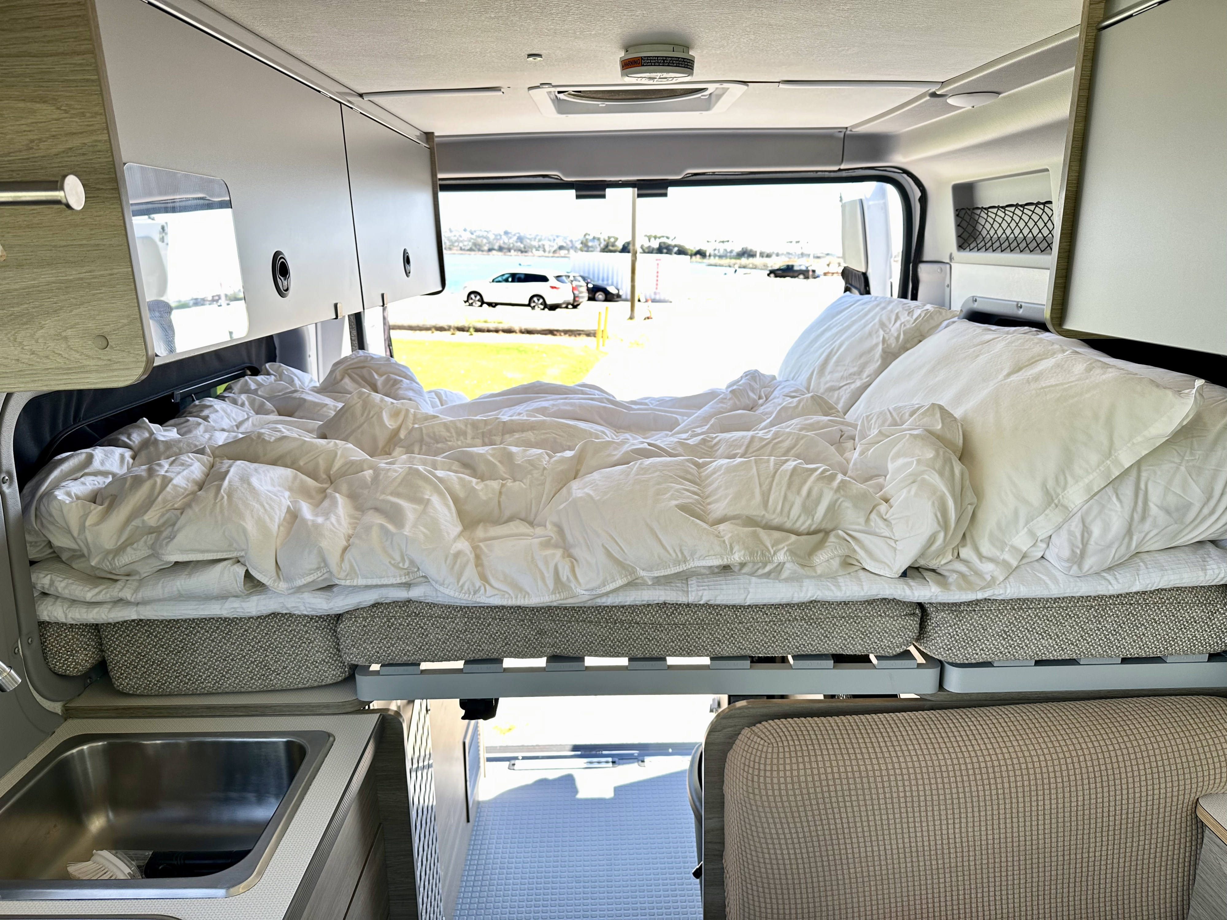 An RV rental camper van interior shows a cozy, raised bed with white linens, a sink, and scenic outdoor views.. Winnebago Solis Pocket 2022