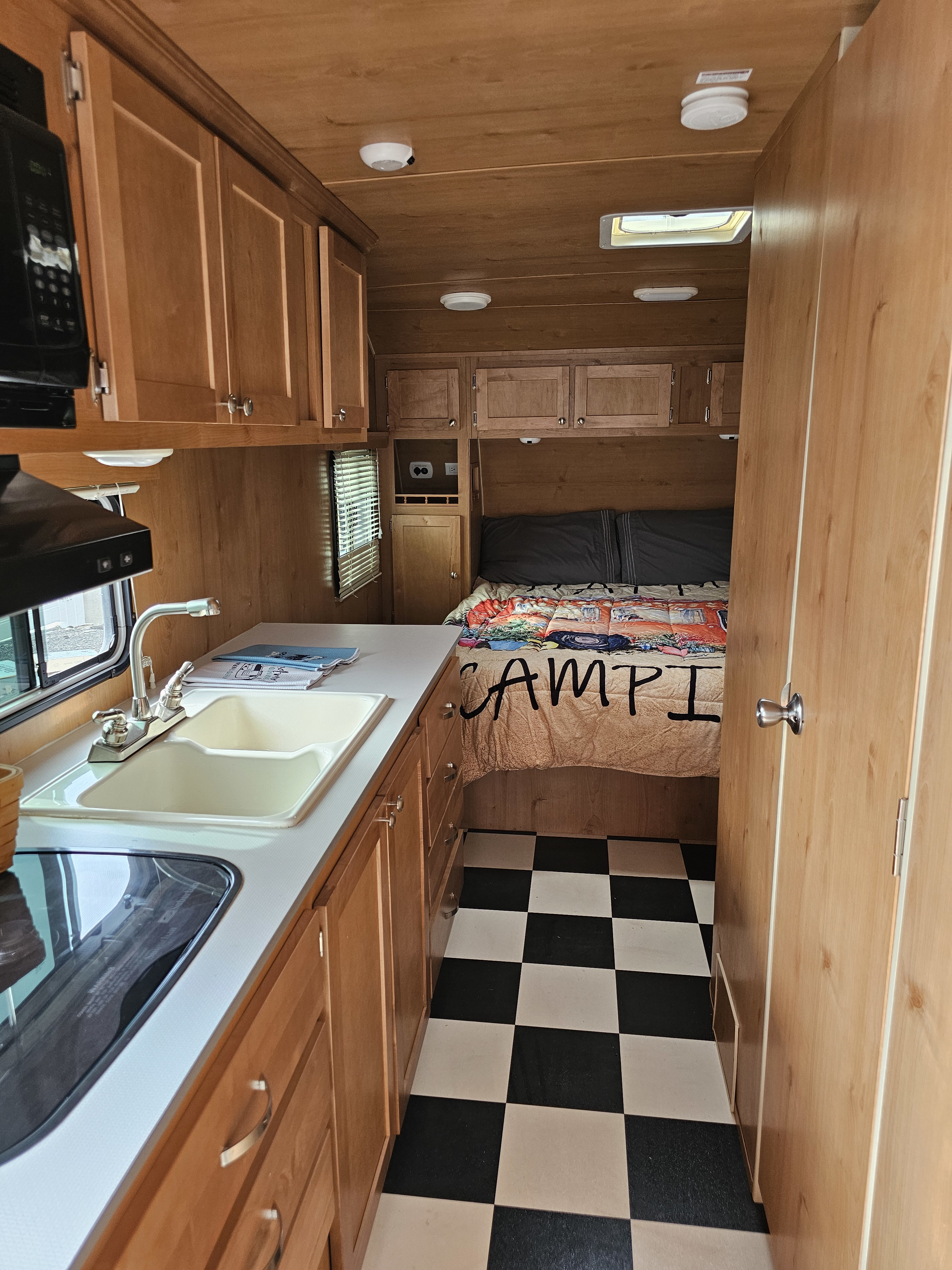 Cozy RV rental interior featuring a compact kitchen, bed, wooden cabinets, and a charming checkered floor.. Riverside RV 195 White Water 2016