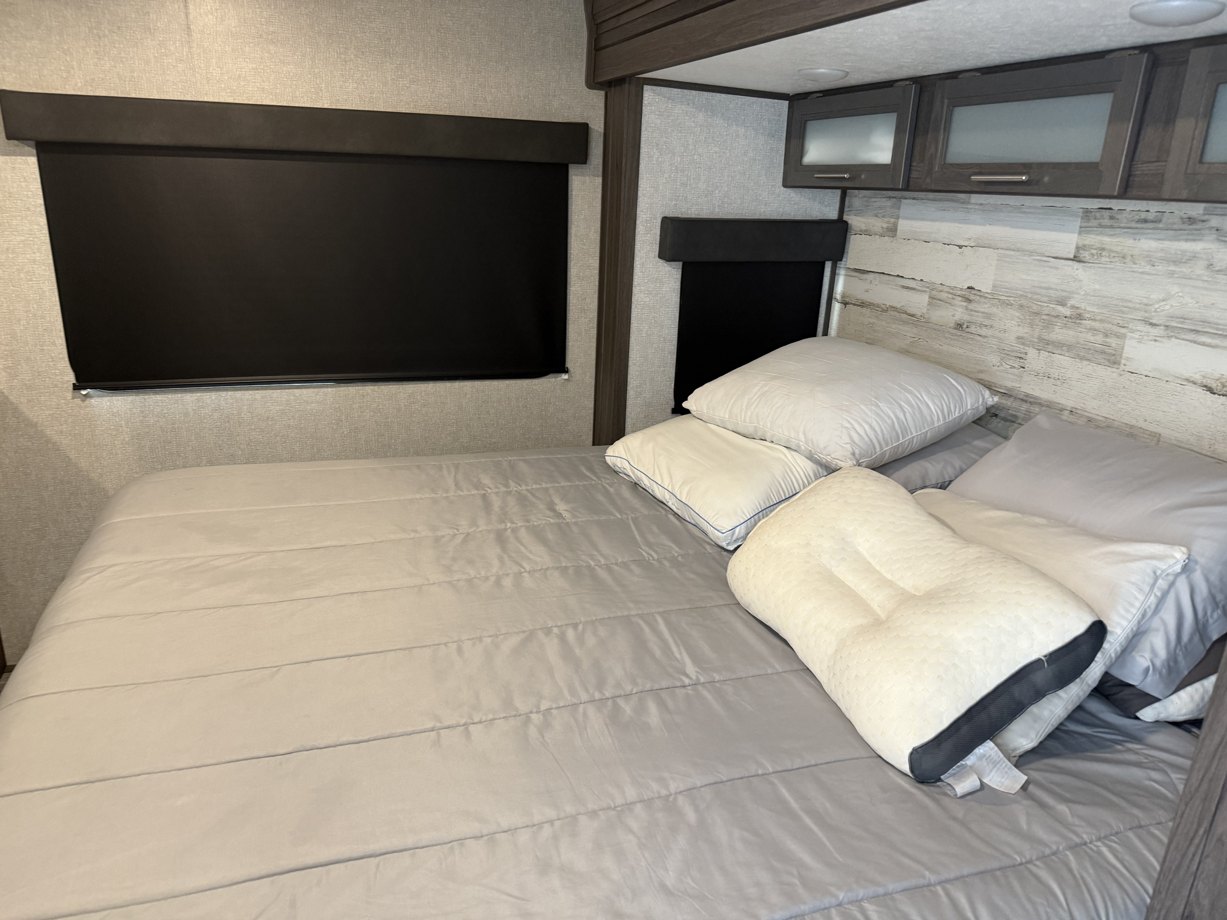 RV rental bedroom with grey bedding, various pillows, blackout shades, a wood-accented headboard, and storage cabinets.. Dutchmen Kodiak Ultimate 2021